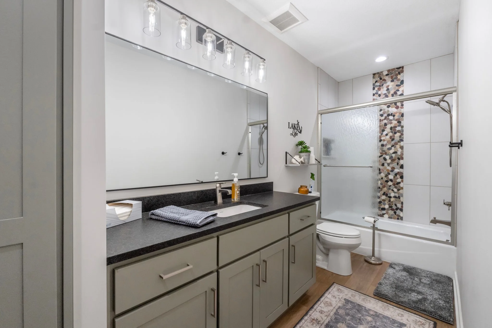 A modern bathroom with a large mirror, vanity with a black countertop, towel, soap dispenser, and decorative items. The bathroom features a toilet, a shower with a frosted glass door, and a combination of white and mosaic tiles. There's a small shelf
