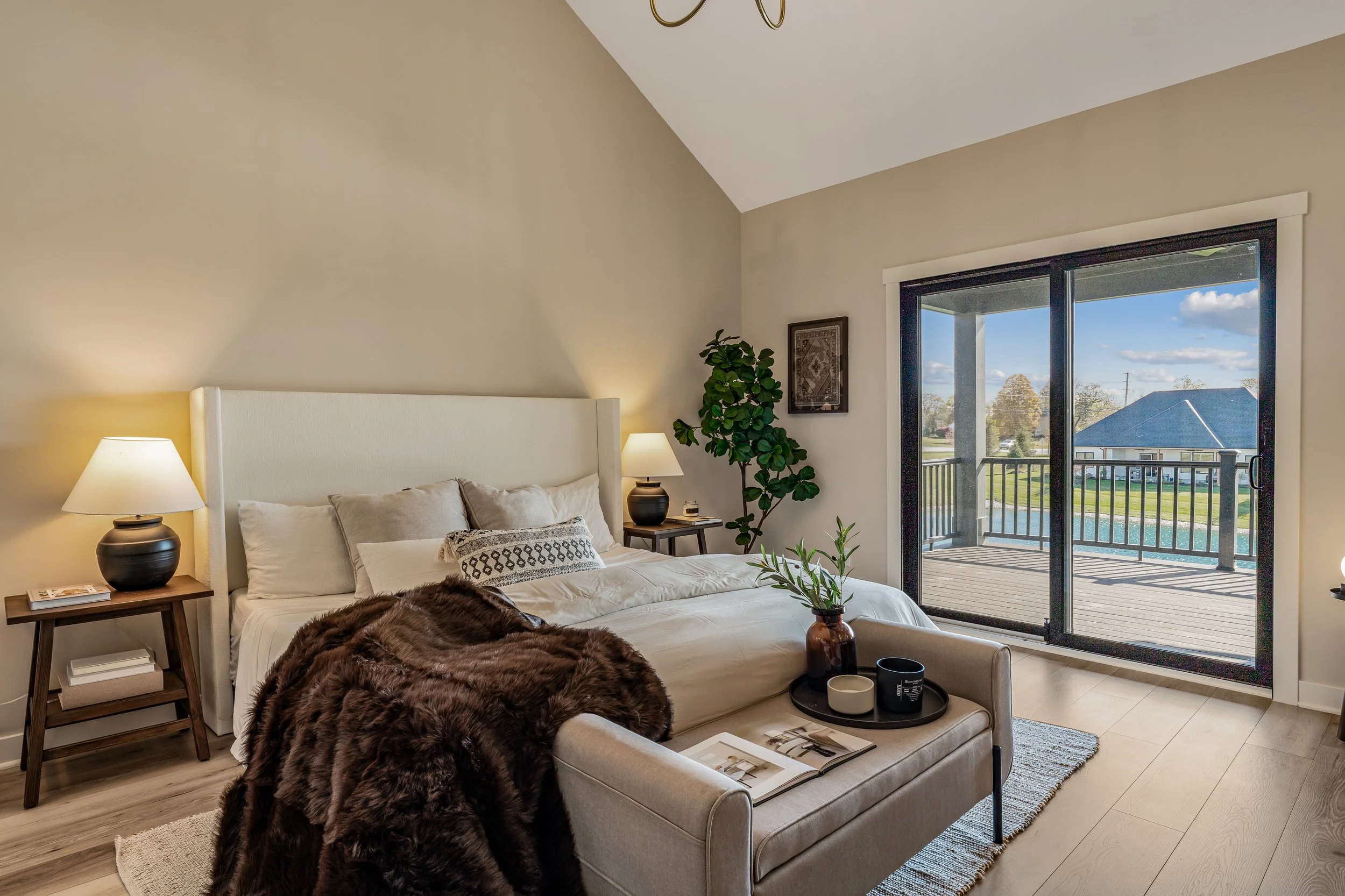 Bright bedroom with a large bed, beige bedding, fur throw, nightstands with lamps, sliding glass door to balcony, wooden flooring, and outdoor view of houses and trees.