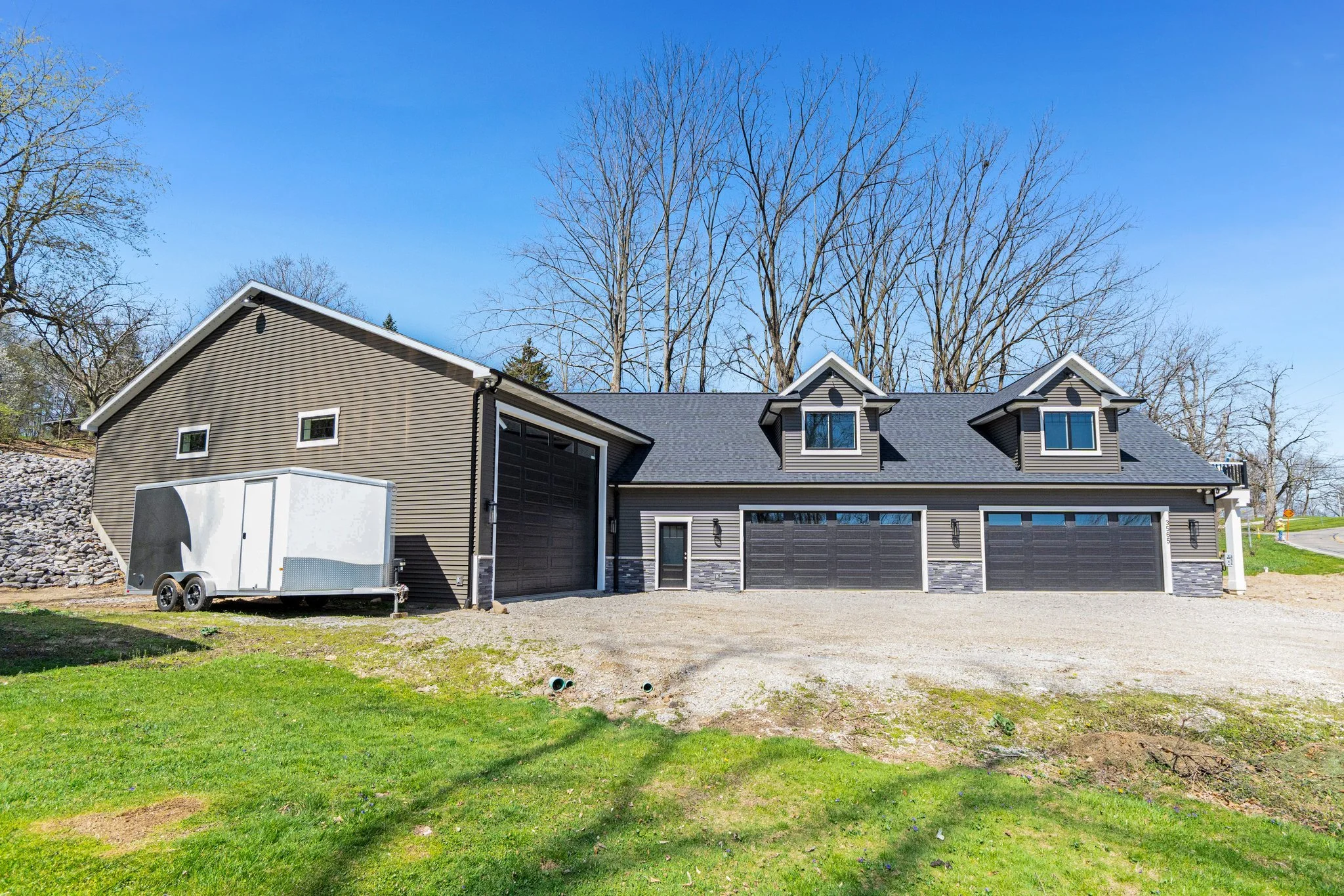 Modern house with gray siding, black garage doors, dormer windows, and a small door, set against a backdrop of leafless trees and a clear blue sky.