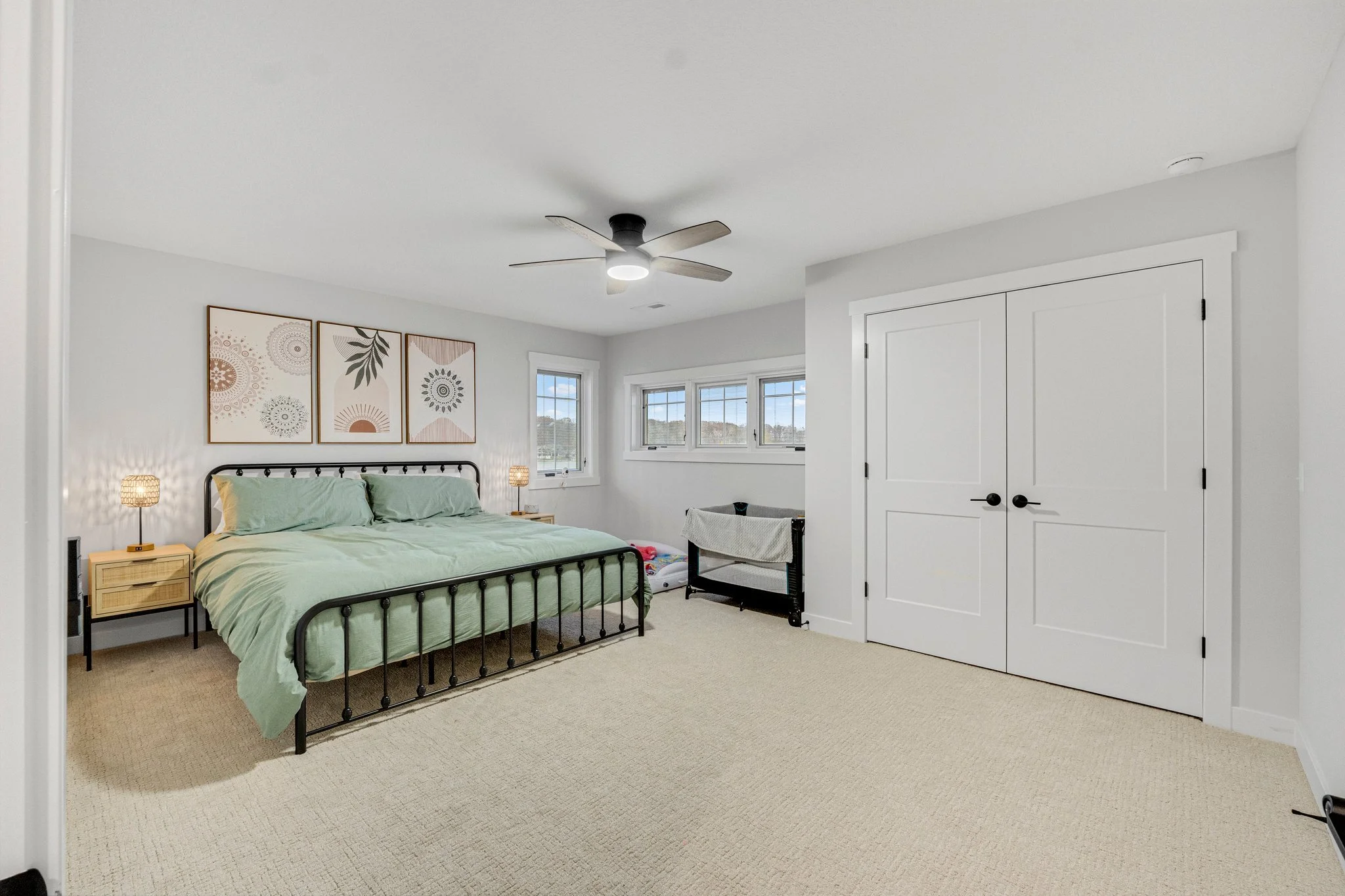 A bedroom with a black metal bed frame, light green bedding, and matching pillows. On each side of the bed are small nightstands with woven lamps. Above the bed are three abstract art pieces. The room has multiple windows allowing natural light. Ther