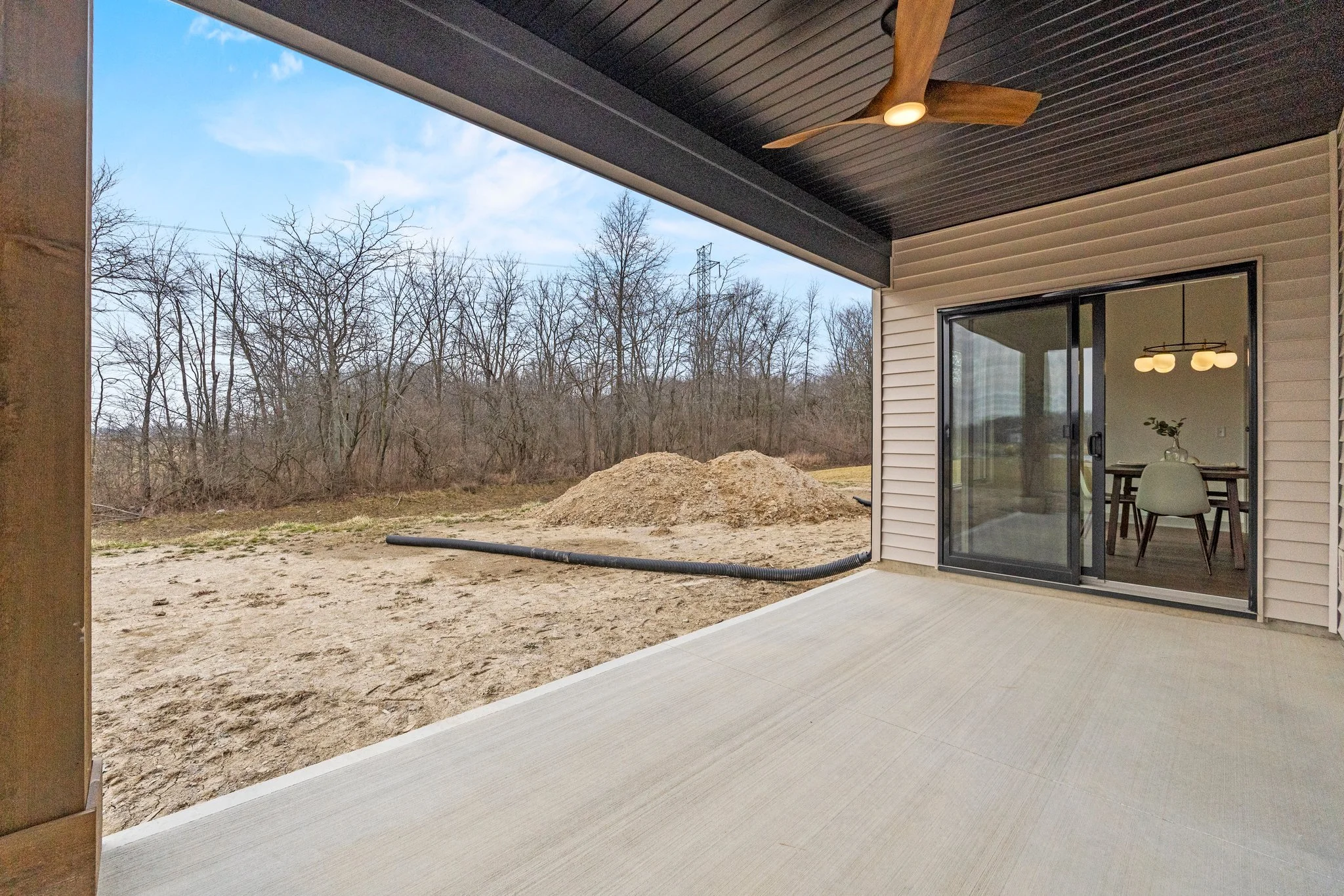 A house with a covered patio area looking out onto a dirt yard with a pile of dirt and a black drainage pipe. The house has beige siding, a sliding glass door, and a black ceiling fan with wooden blades. Inside the house, a modern light fixture and a