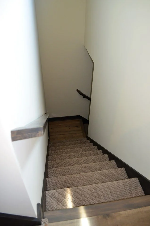 Indoor staircase with beige carpet and wooden handrail, viewed from the top looking downward.