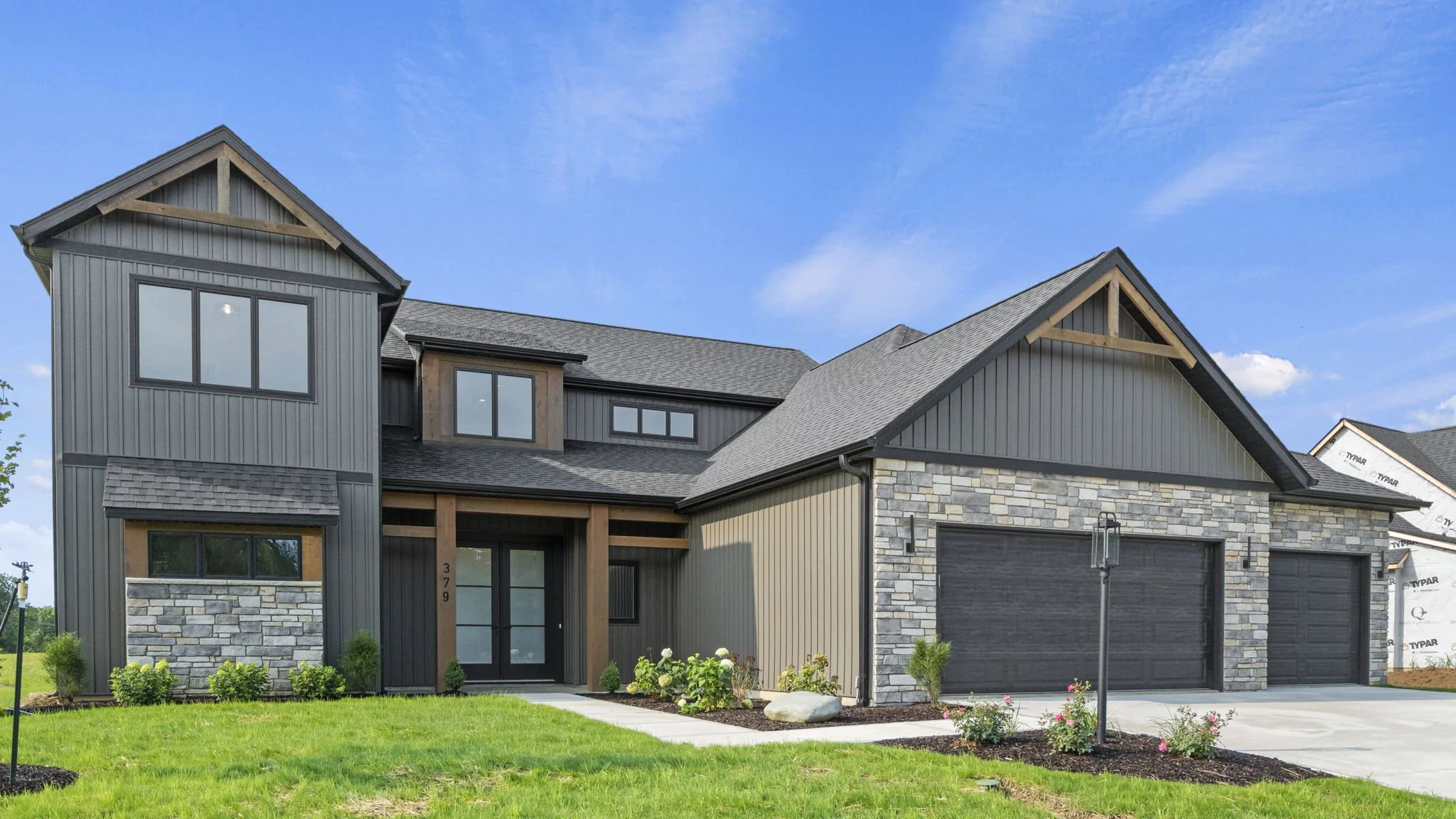 New modern house with stone, metal, and wood exterior in a suburban neighborhood, green lawn, and a blue sky.