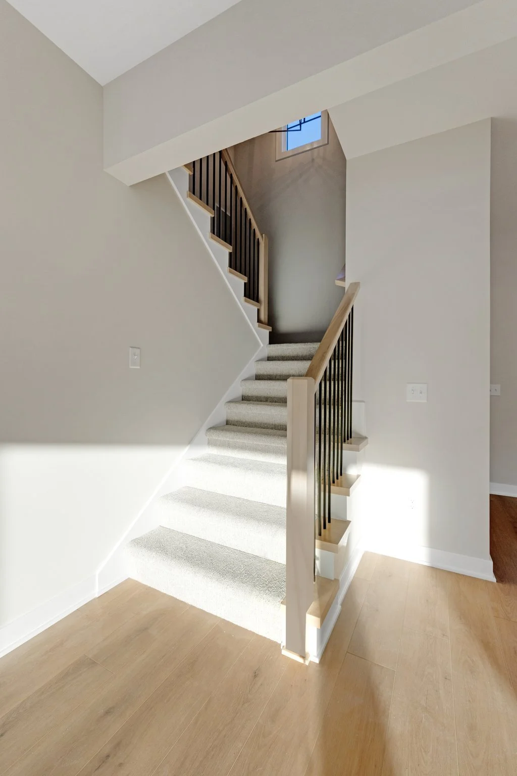 Interior view of a staircase with light-colored carpet, wooden handrails, and black balusters, leading up to an upper floor with a small window letting in natural light.