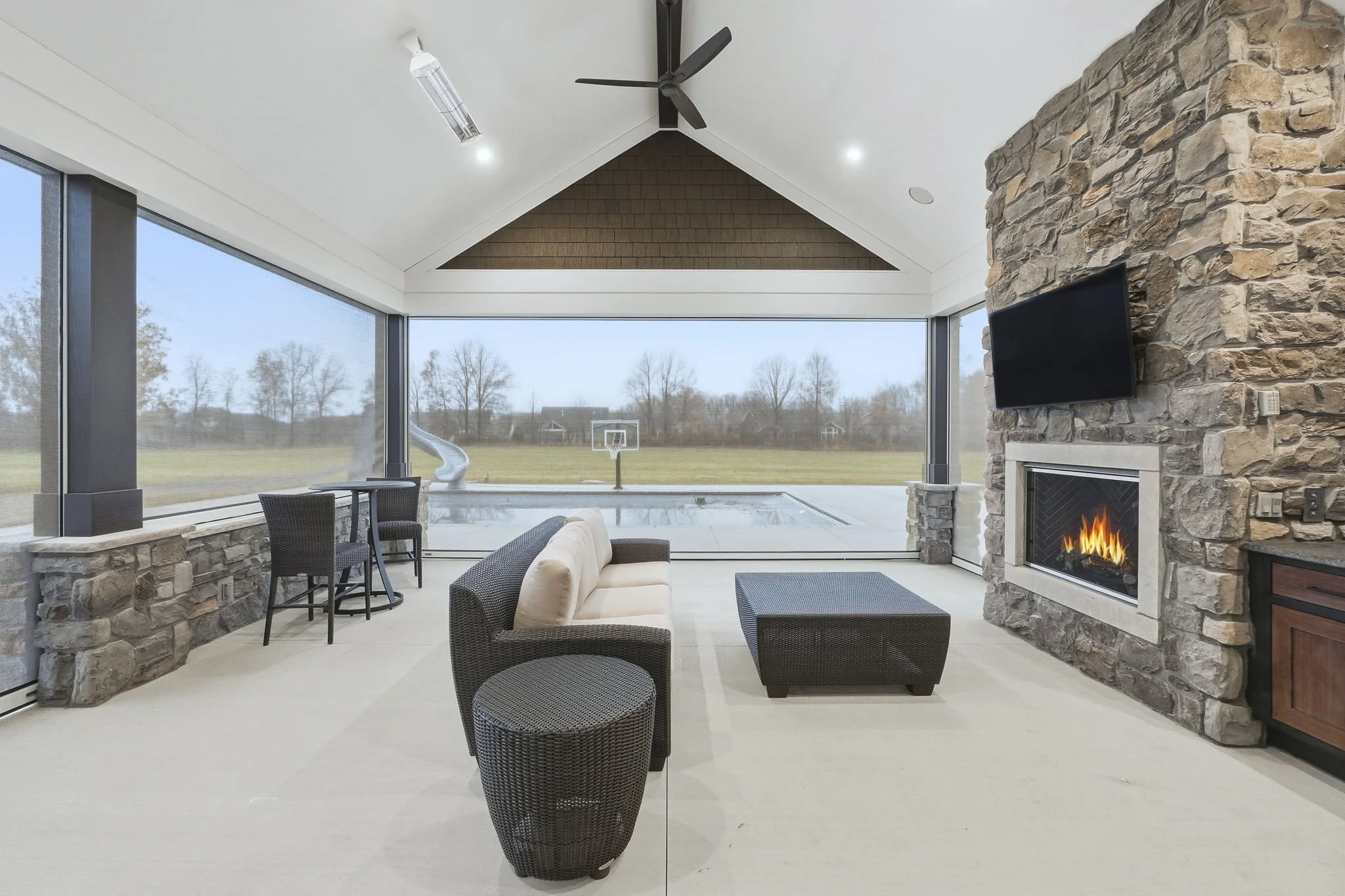 A modern sunroom with large windows overlooking a backyard with a pool and a basketball hoop. The room has a stone fireplace, a mounted TV, a ceiling fan, and outdoor seating including a sofa, a round table with chairs, and an ottoman.