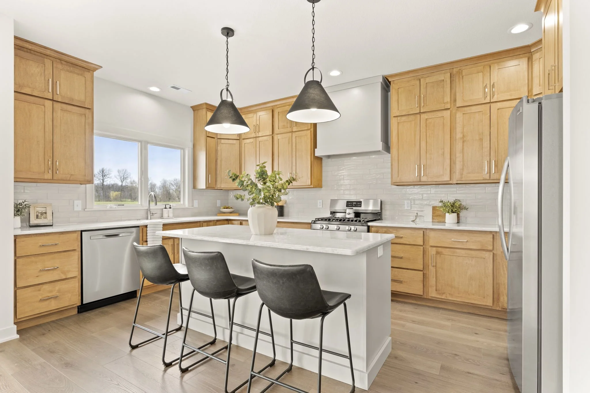 Modern kitchen with wooden cabinets, white countertop, stainless steel appliances, kitchen island with three black chairs, large window with a view of trees, potted plant on the island, and pendant lights.