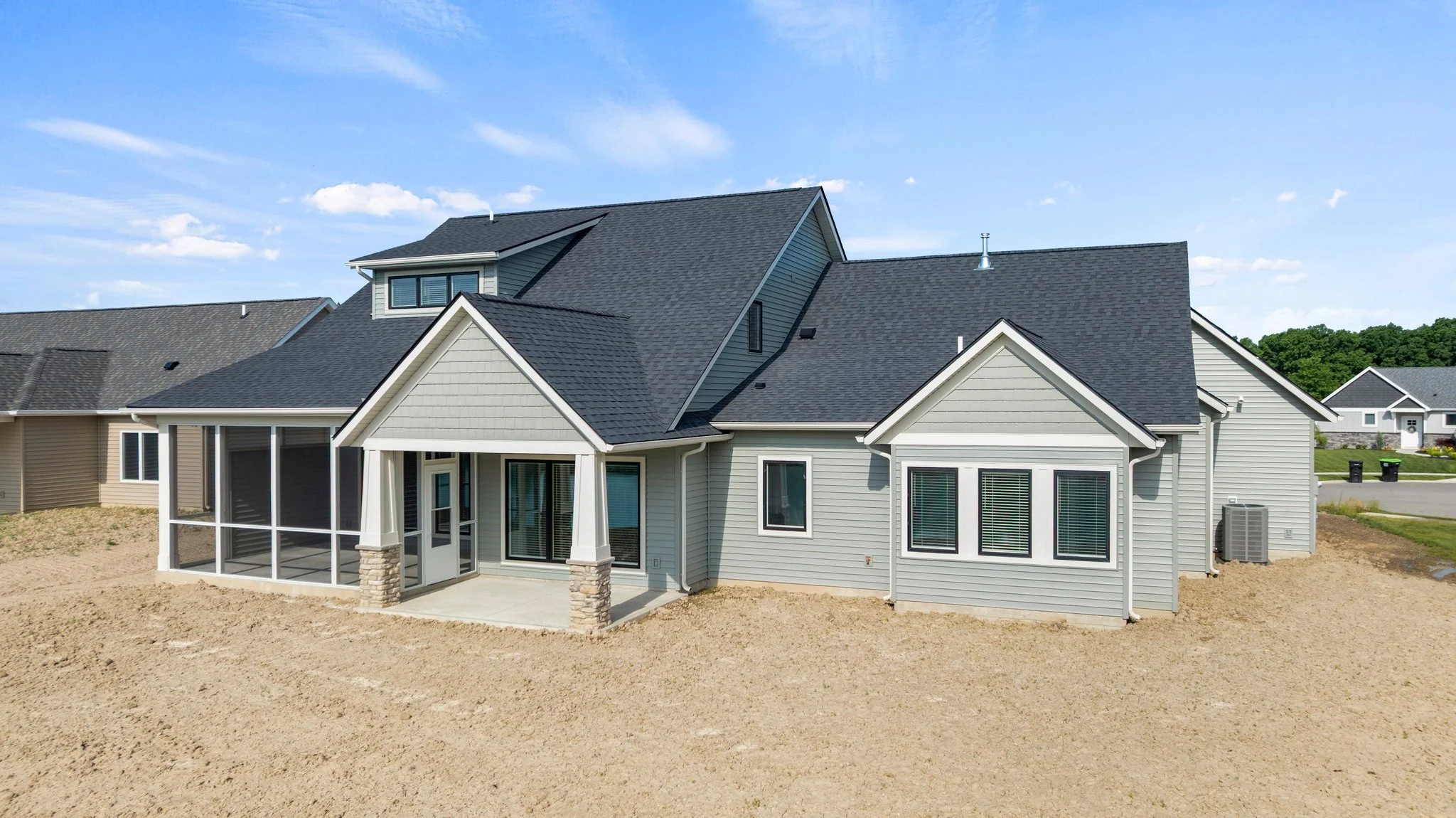 Newly constructed two-story house with gray siding, black roof, multiple windows, and a screened porch in the backyard, on a dirt lot in a suburban neighborhood.