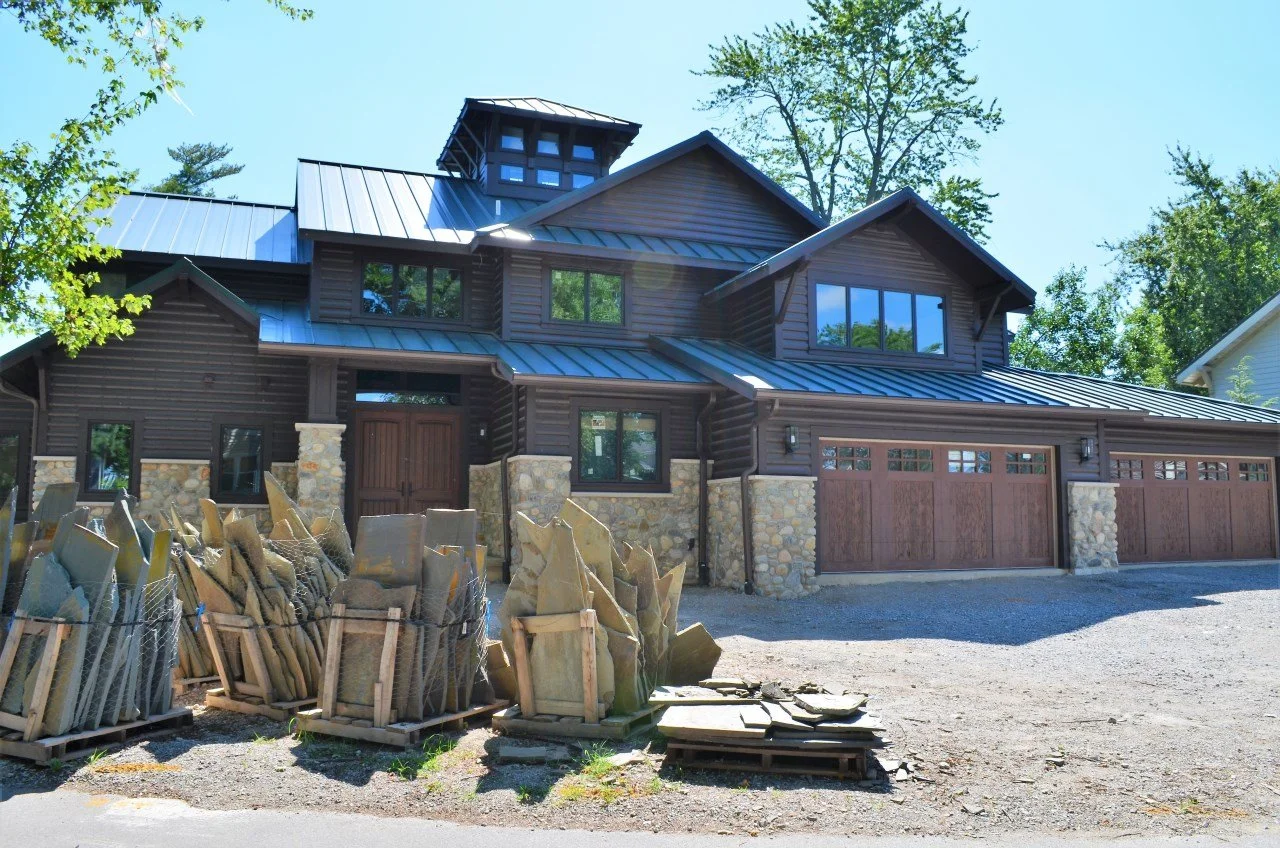 A new modern house with a dark wood exterior, stone accents, and large garage doors, surrounded by trees, with construction materials in the front yard.
