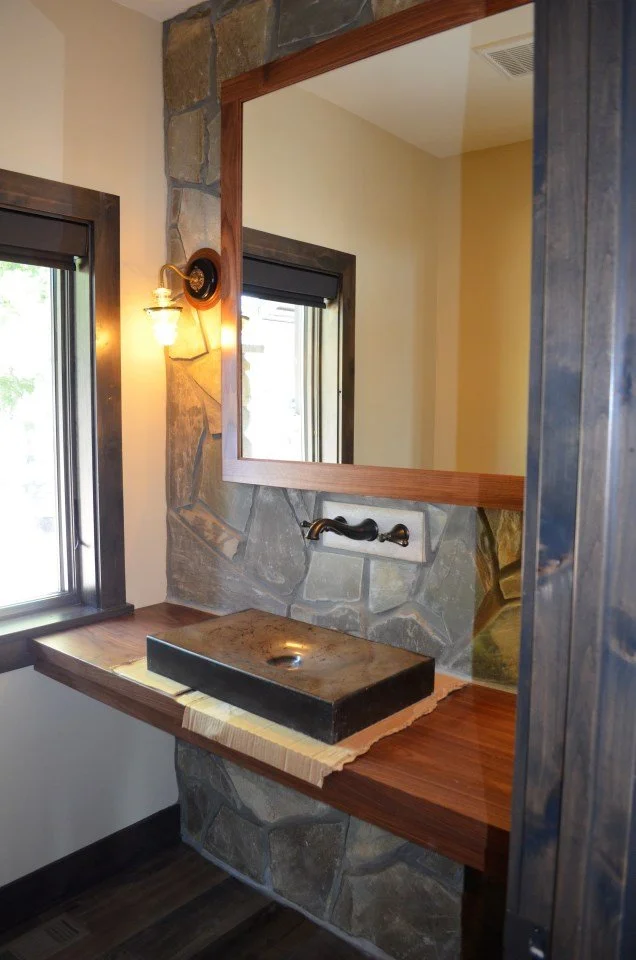 Close-up of a rustic bathroom vanity with a stone wall and a large mirror. The vanity features a dark stone basin, a vintage-style faucet, and wooden countertops.