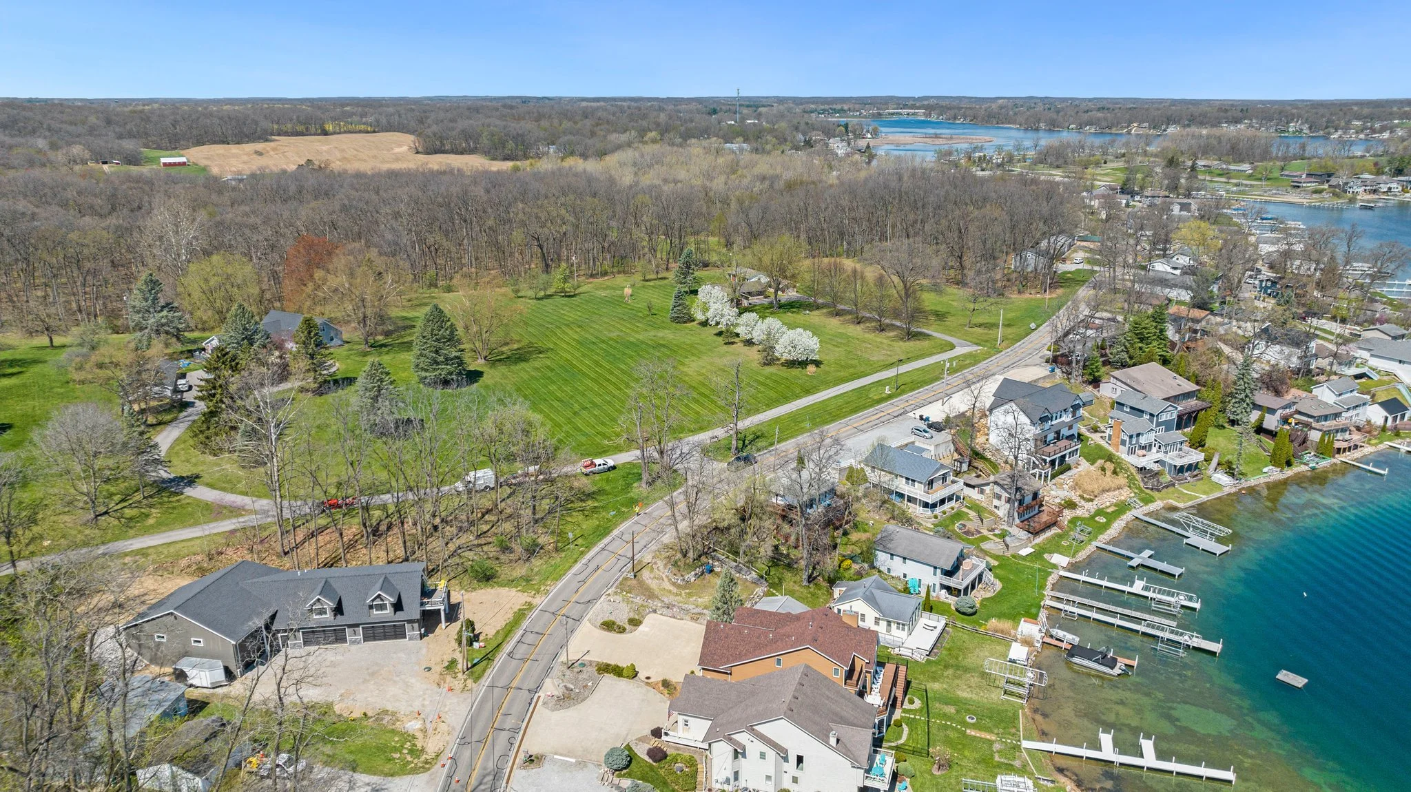 Aerial view of a lakeside residential neighborhood with houses, docks, and boats, adjacent to a large green park with trees and a road winding through the area.