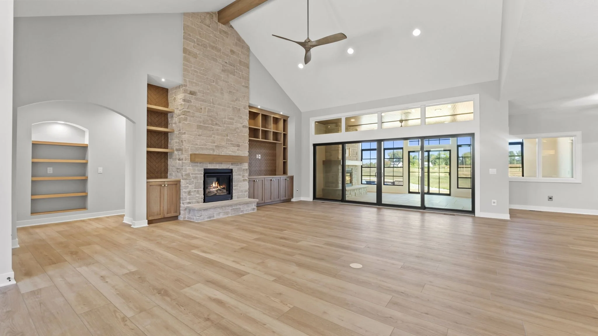 Empty living room with a stone fireplace, wooden built-in shelves and cabinets, large sliding glass doors, and light-colored hardwood floors.