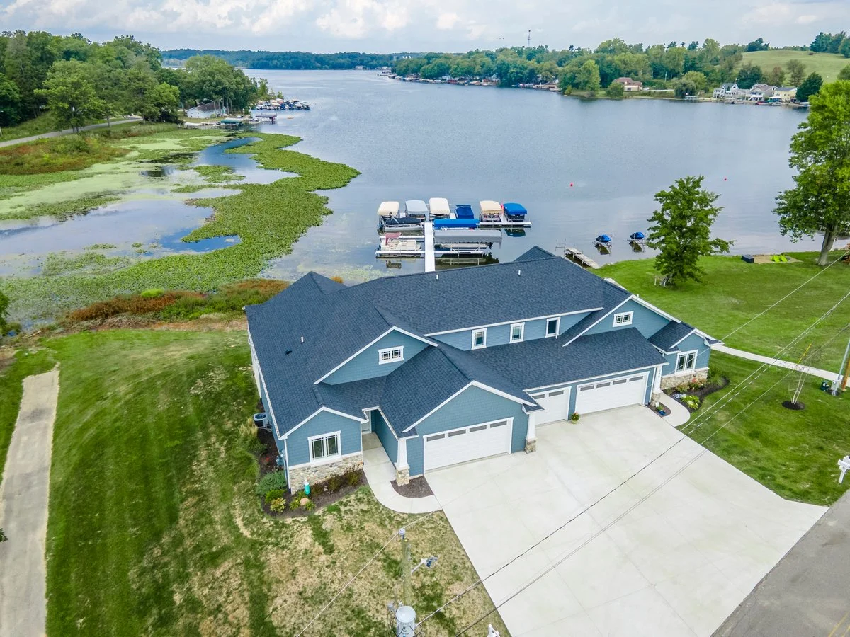 Aerial view of a blue house with a large driveway, situated next to a lake with boats docked at a marina.