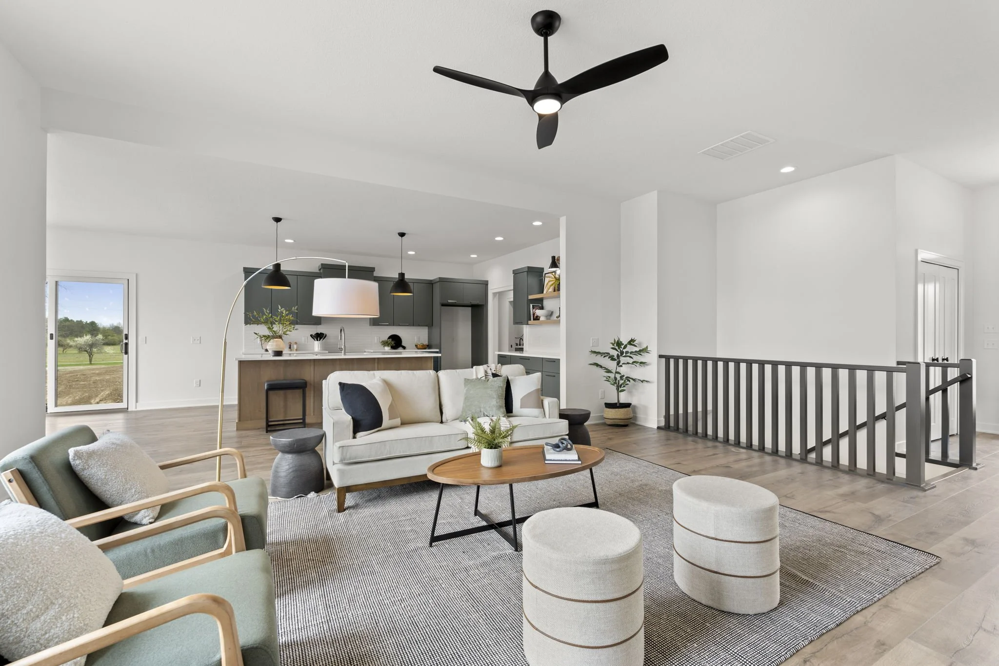 Open living room and kitchen with white walls, high ceiling, black ceiling fan, and light wood flooring. Seating includes a beige sofa with black and green pillows, two green armchairs, and two beige ottomans. Coffee table with a plant and book, side