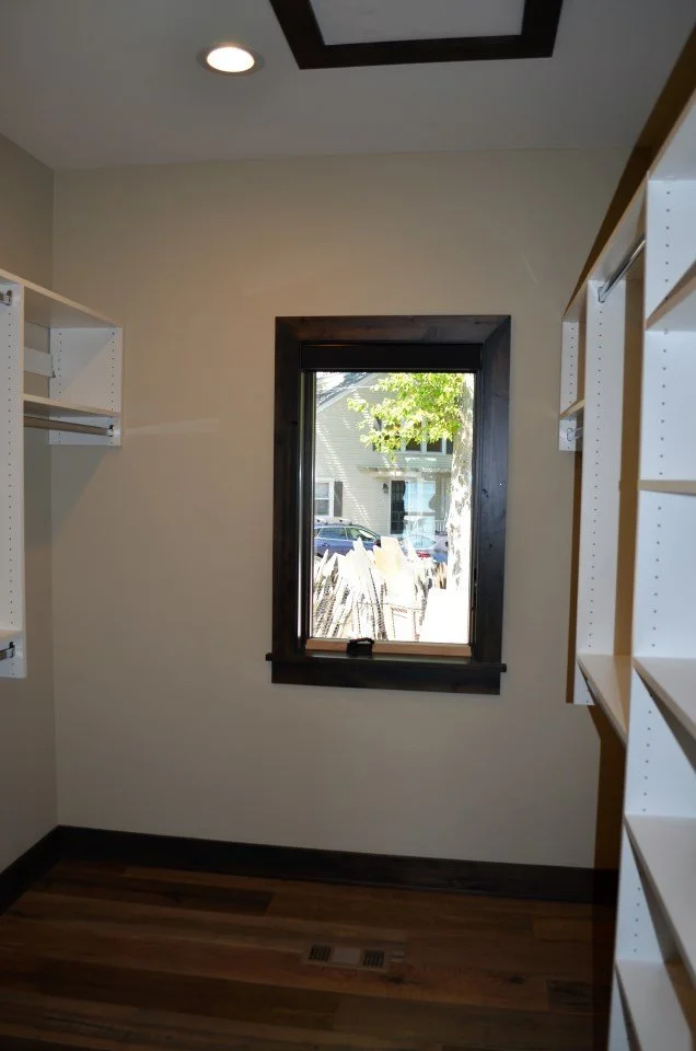 Small room with a single window, wooden trim, and a hardwood floor. Shelves are on the walls, and a vent is on the floor. Outside the window, trees and a house are visible.
