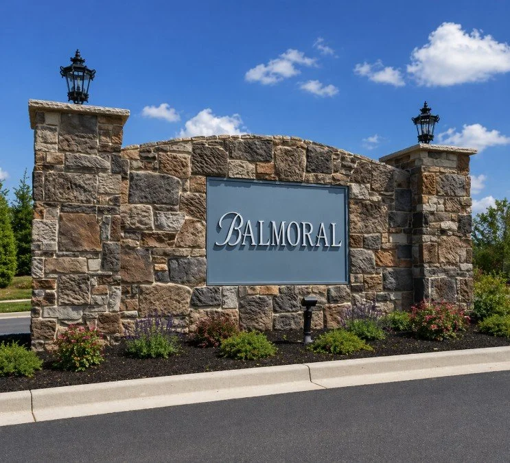 Stone entrance sign with the word 'Balmoral' and decorative lanterns on top, surrounded by landscaped plants.