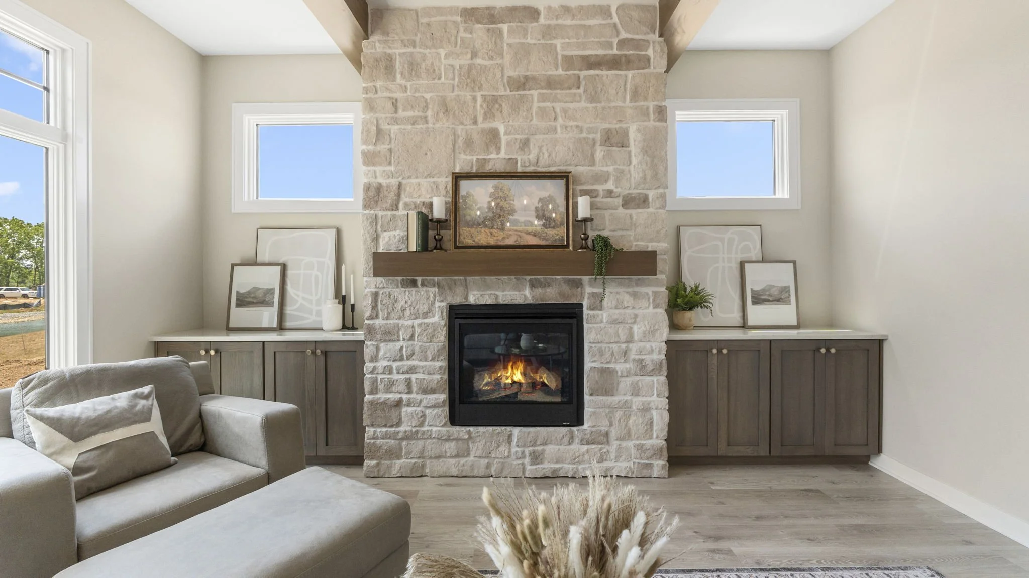 Living room with a stone fireplace, wooden mantel, and built-in cabinets on either side. Framed artwork and decorative items are displayed on the mantel and cabinets, with windows above and beside the fireplace. A beige sofa with pillows is partially