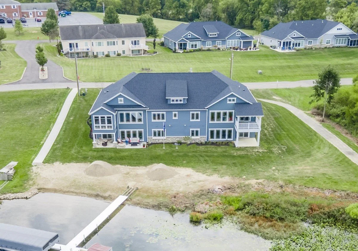 A large blue house on a grassy waterfront lot, with multiple balconies, patio furniture, and a boat dock leading to a small lake or pond.