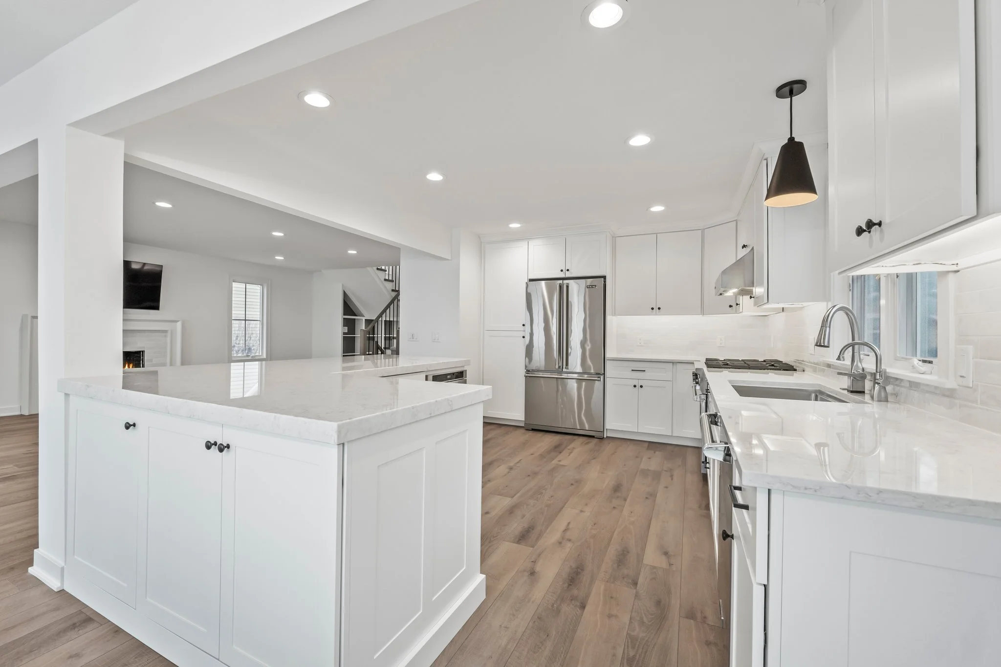 Modern white kitchen with marble countertops, stainless steel refrigerator, and wood flooring.
