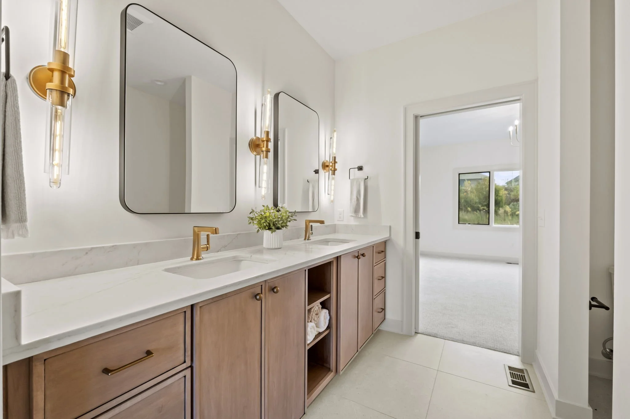 Modern bathroom with dual sinks, wooden cabinets, marble countertop, and two rectangular mirrors, illuminated by vertical wall sconces, with a view into a bright, empty bedroom with a window.