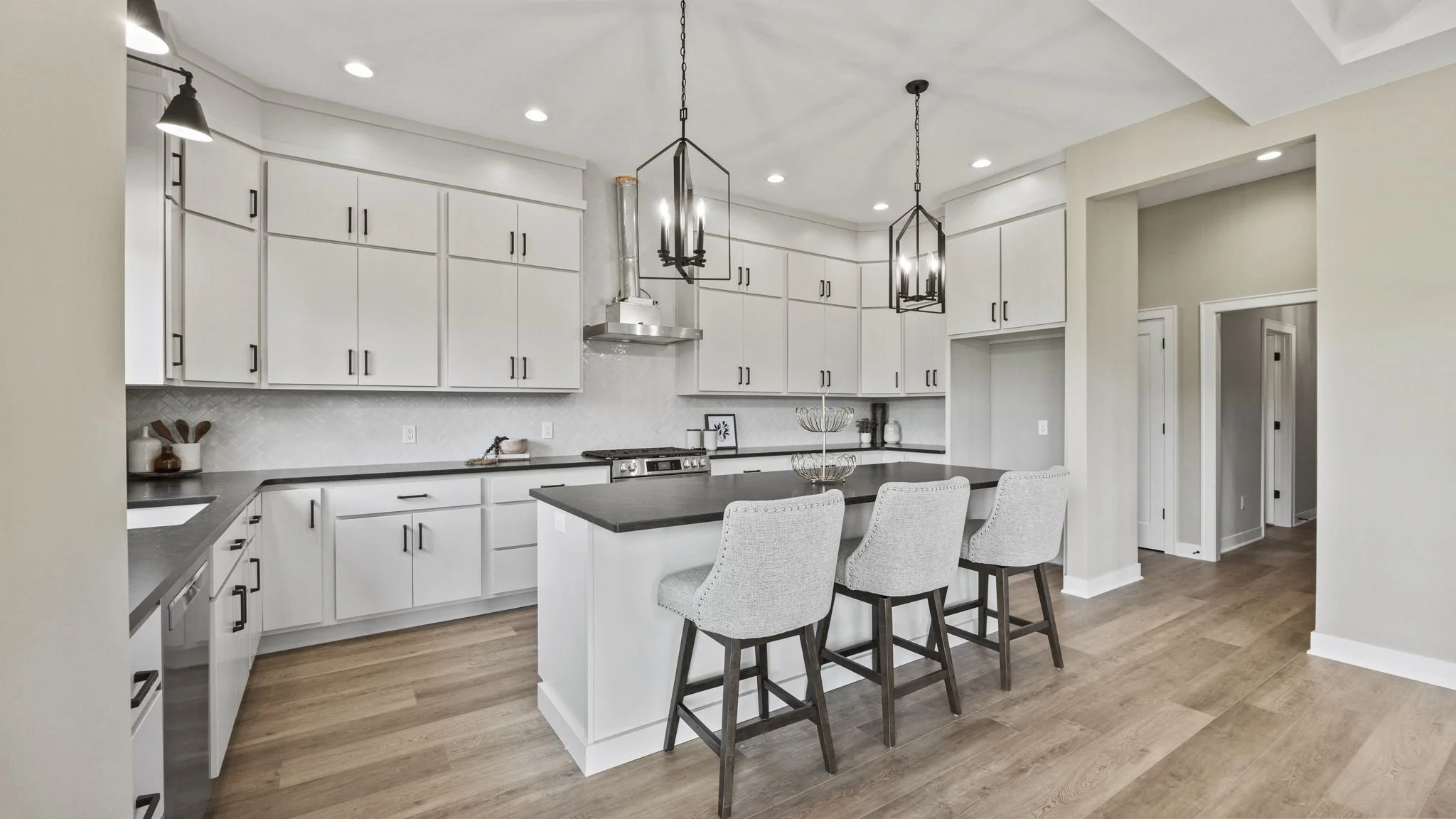 Modern white kitchen with black countertops, an island with three light gray chairs, and black pendant and ceiling lights, with wood flooring and a hallway leading to other rooms.