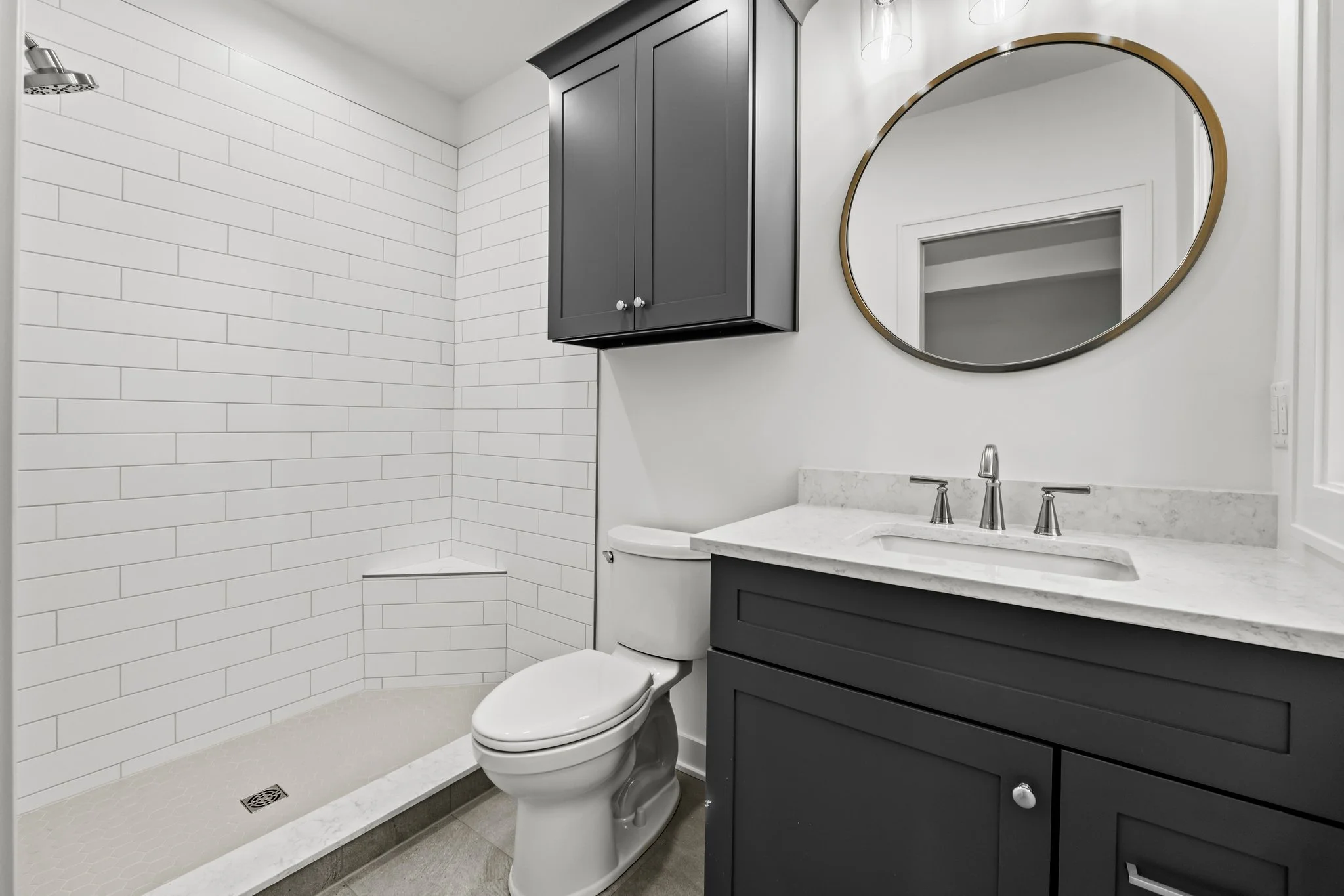 Modern bathroom with a walk-in shower featuring white subway tiles, a white toilet, and a dark gray vanity with a marble countertop and an oval mirror.