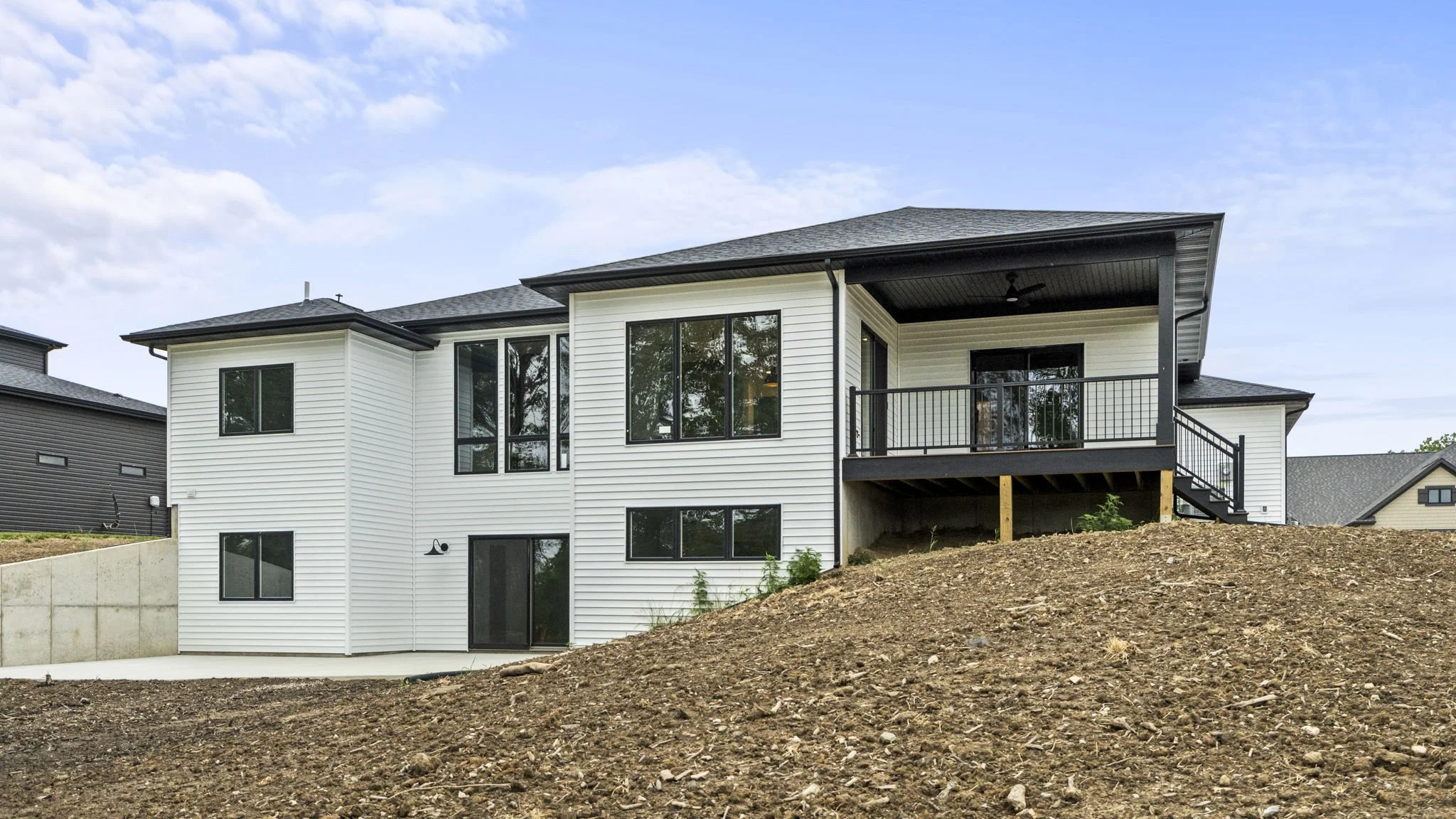 Modern two-story house with white siding, black window frames, and a black roof. The house has a covered balcony with a black railing and a staircase leading down to the yard. The yard is terraced with dirt and some small plants.