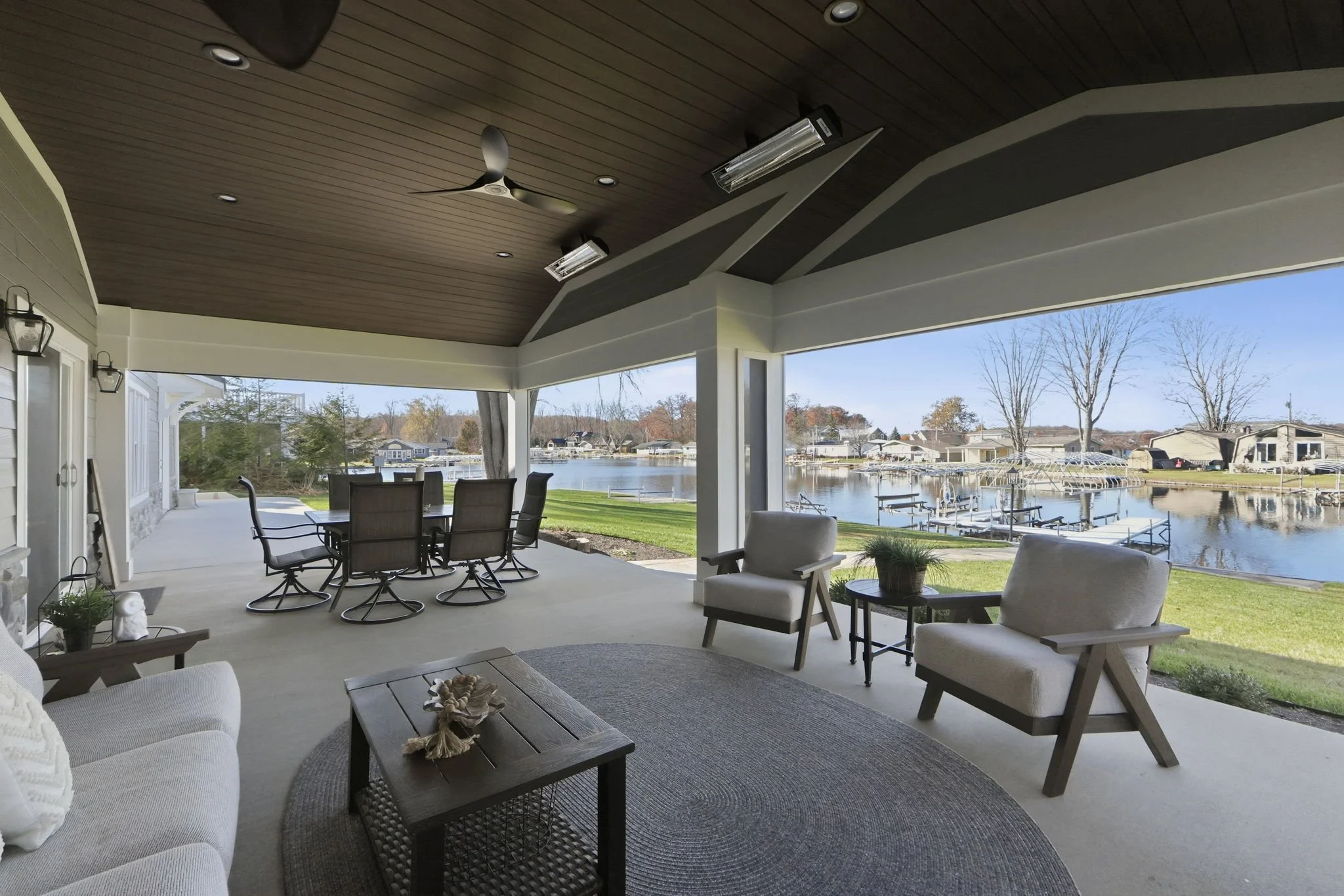 Covered patio with outdoor furniture overlooking a waterway with docks and houses, featuring a ceiling fan, ceiling lights, and a view of trees and a blue sky.