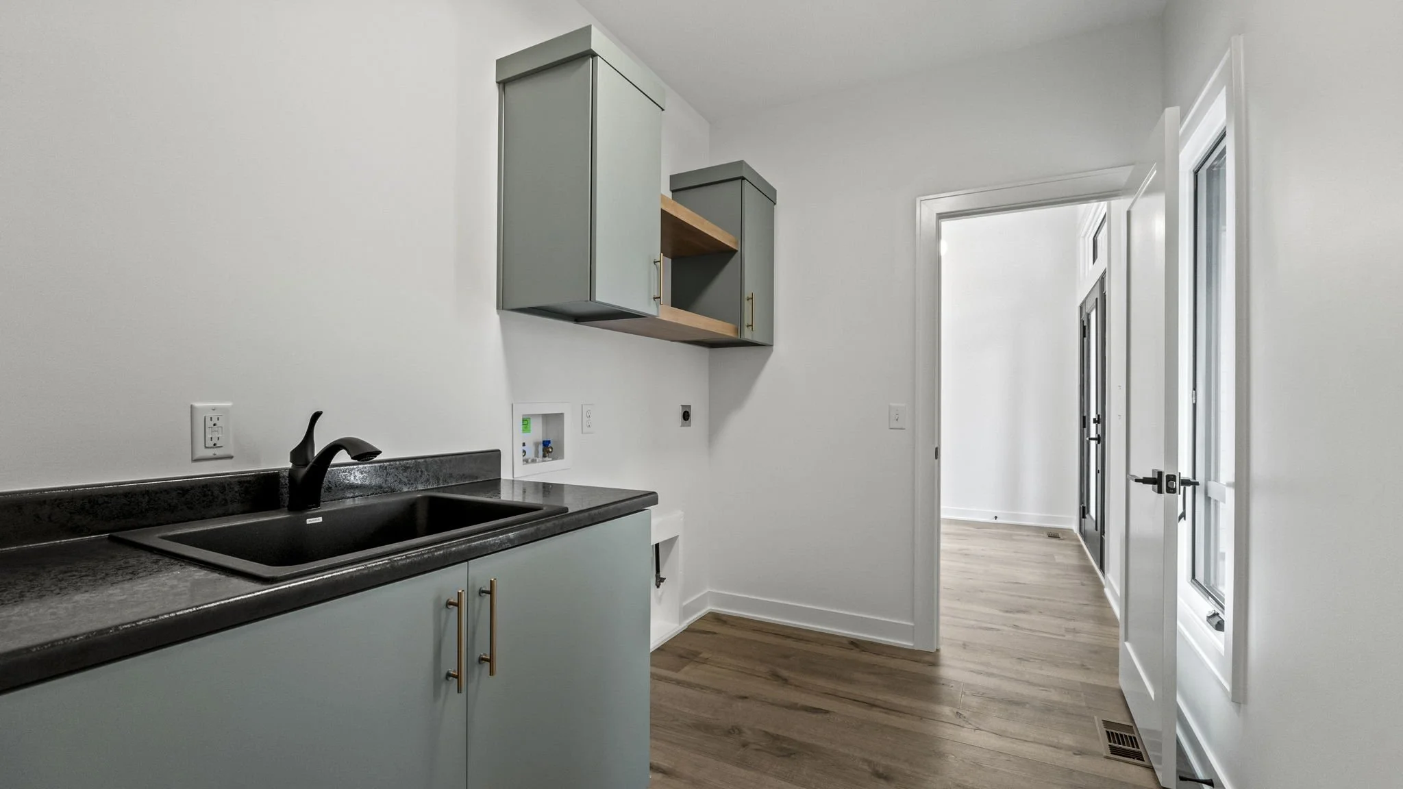 Empty kitchen with black countertop, blue cabinets with gold handles, a black faucet, and wooden floors. There are some wall outlets and unfinished hookups on the wall, with open doors leading to bright rooms.