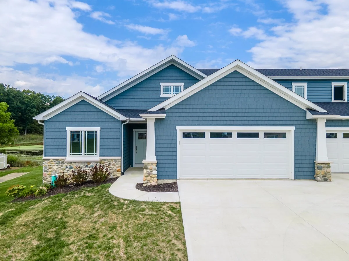 Front view of a blue house with a white garage door, stone accents, and a small front porch. The sky is partly cloudy.