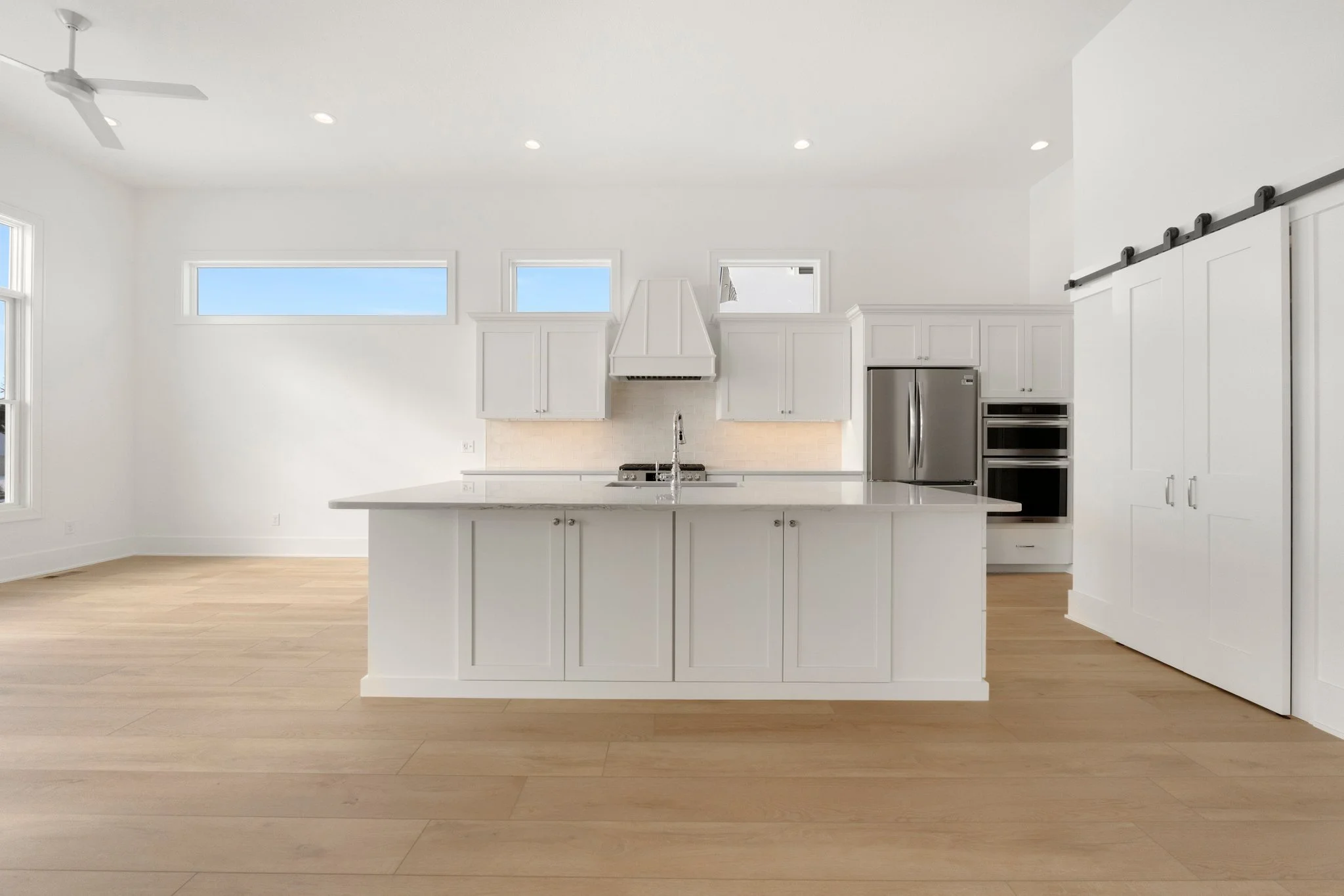 Empty modern kitchen with white cabinets, stainless steel appliances, light wood flooring, and high windows.