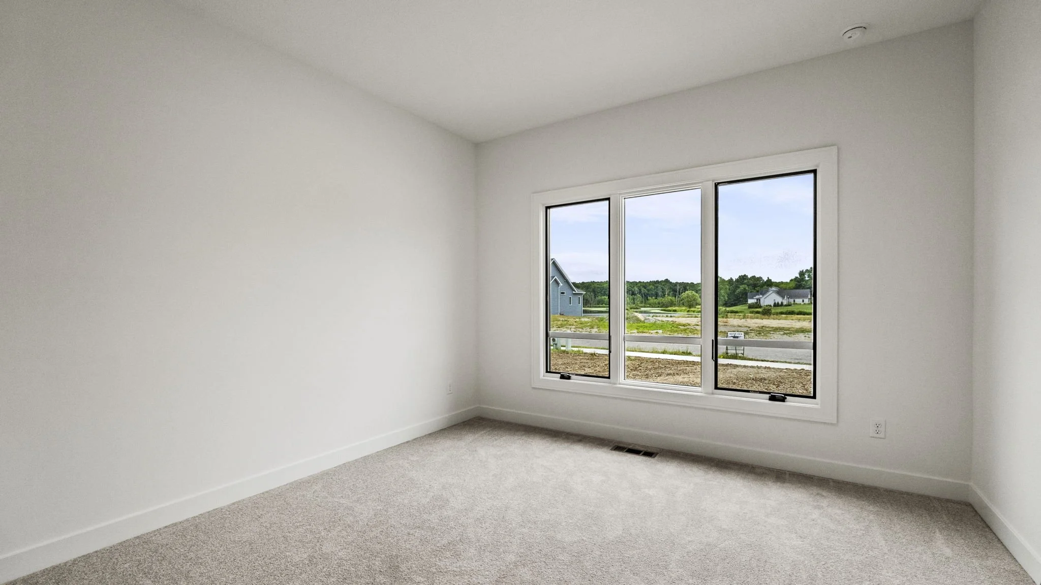 Empty room with white walls, beige carpet, and large window showing a view of residential houses and trees outside.