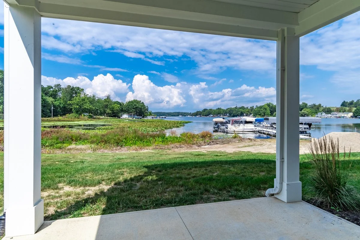 View from a covered porch overlooking a river with boats docked at the shore, green trees, and a partly cloudy sky.