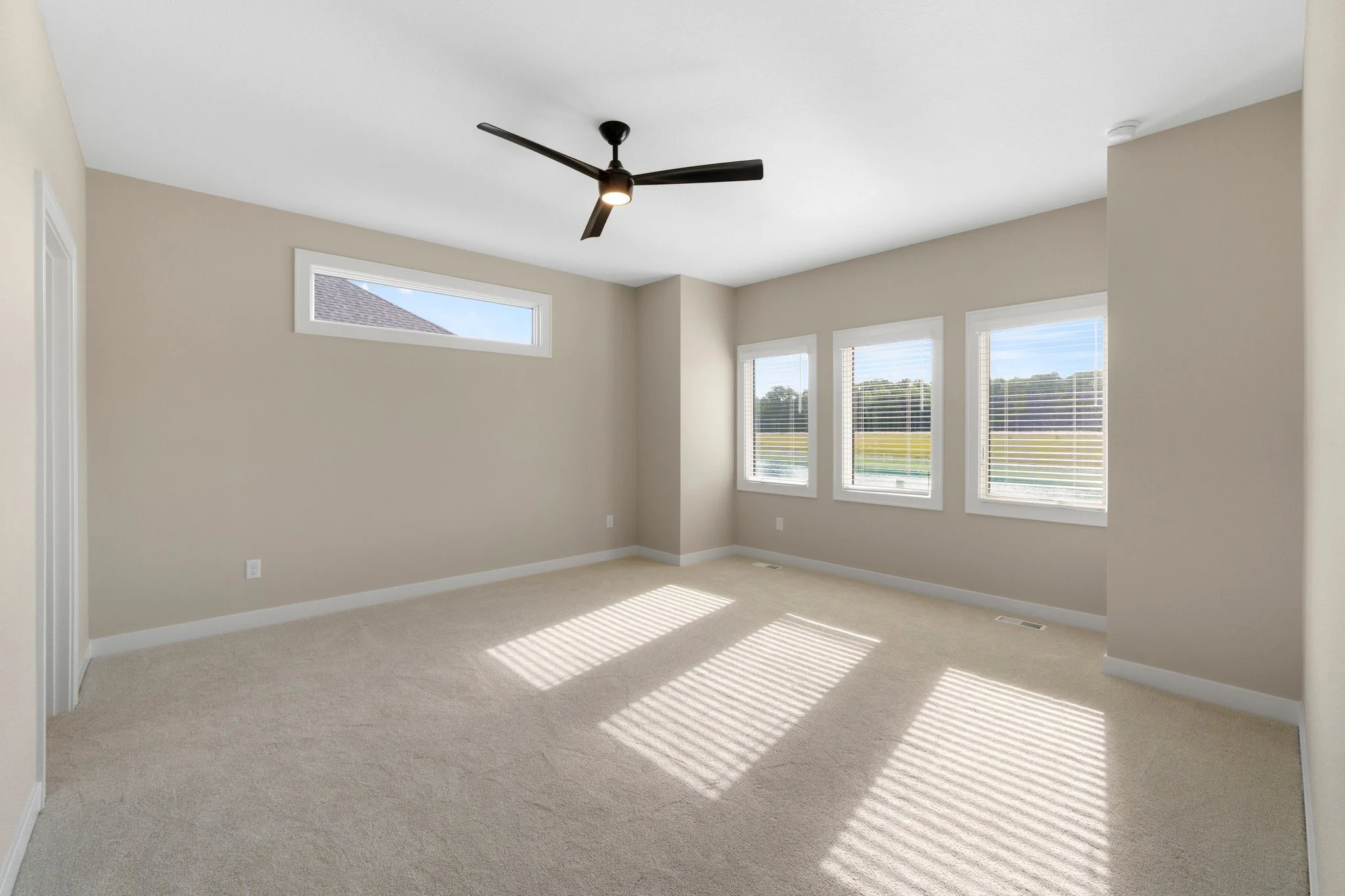 Empty room with beige walls, white trim, a ceiling fan, and three large windows with blinds, sunlight casting shadows on carpeted floor.