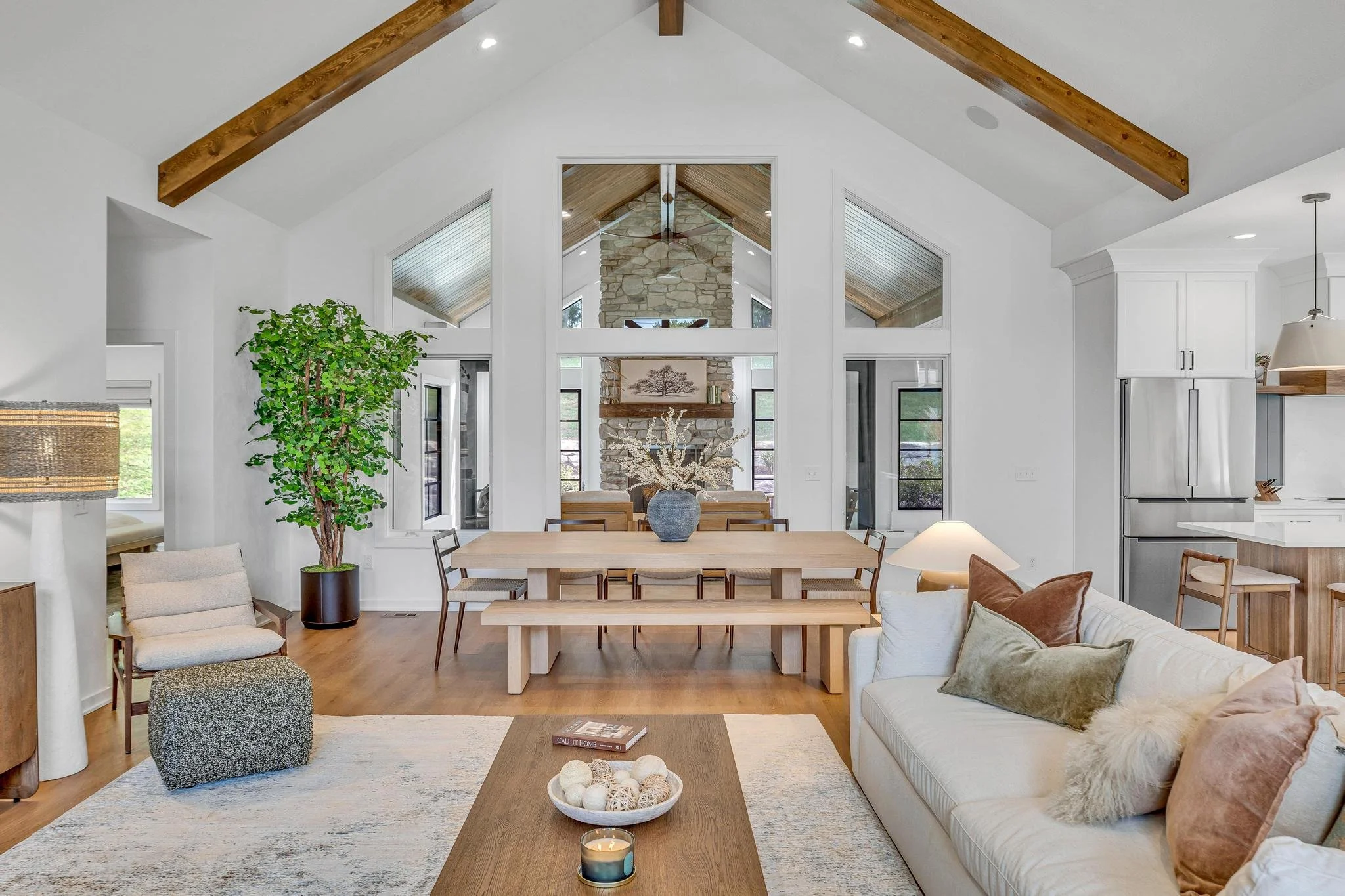 Open floor living room and dining area with vaulted ceiling, exposed wooden beams, and a stone fireplace. Furniture includes a white sofa with decorative pillows, a wooden dining table with benches and chairs, and a standing lamp. Large windows and a