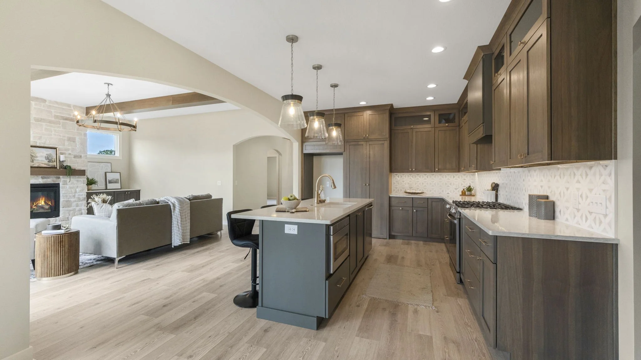 Modern open-concept kitchen and living room with wooden cabinets, a kitchen island with a sink, three pendant lights, and a living area with a fireplace and gray sofa.