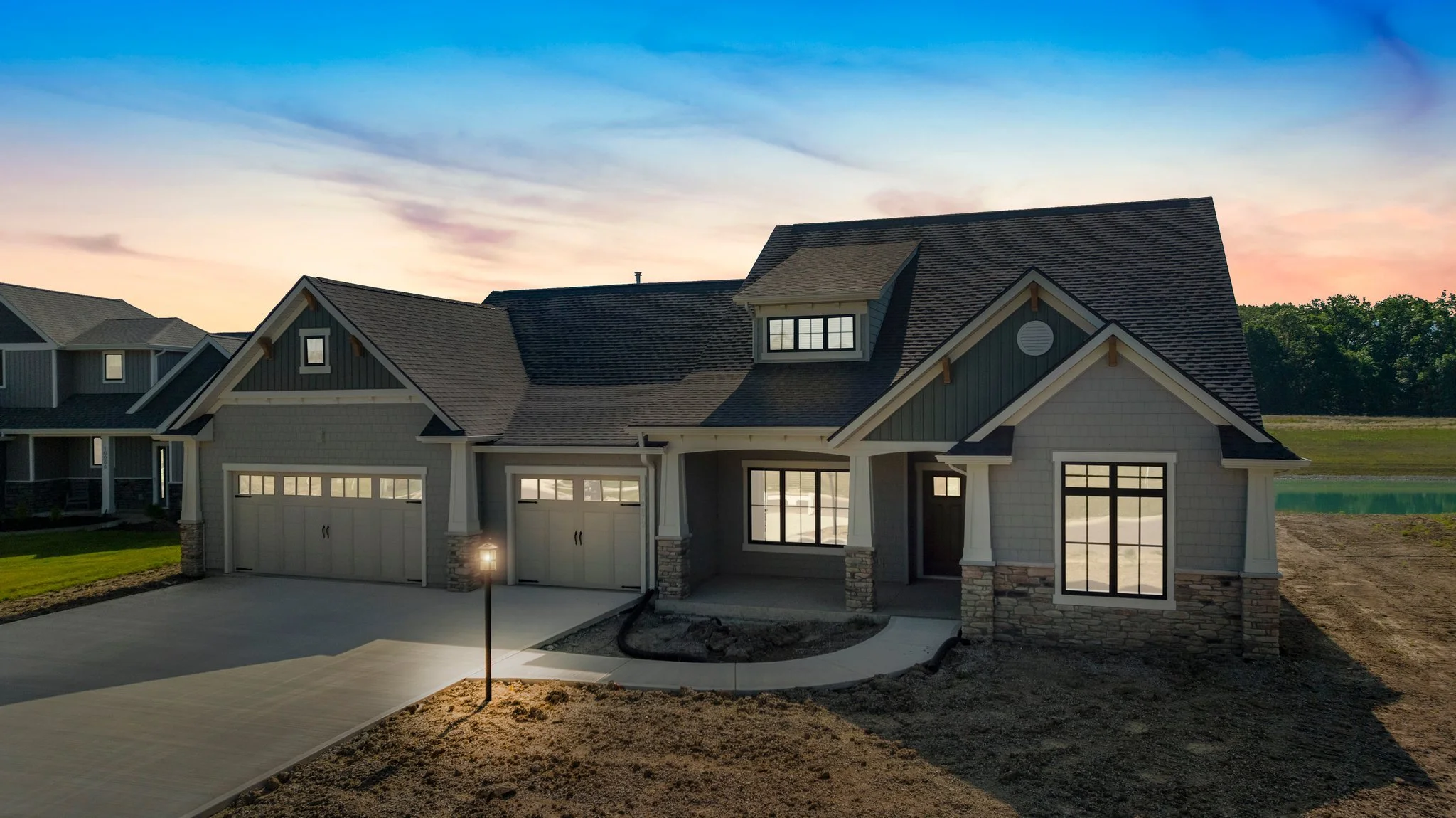 Newly constructed two-story house with gray siding, black roof, and large windows, under a colorful sky at sunset, surrounded by a driveway and yard.