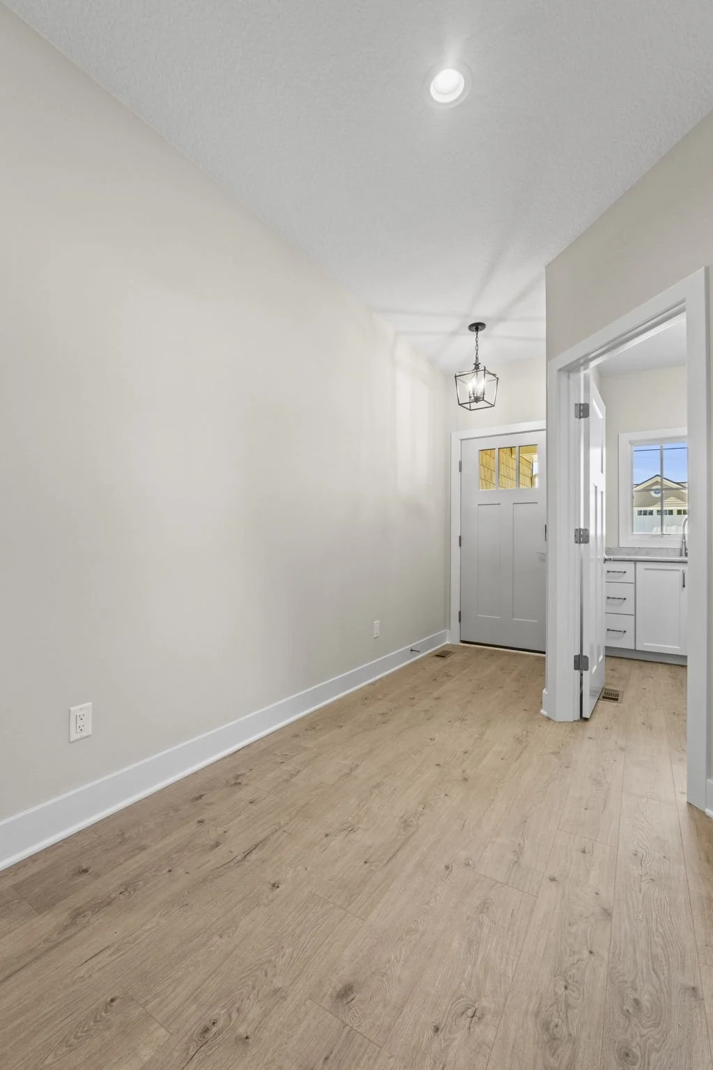 Empty hallway with light wood flooring, white walls, a front door, and a view into the kitchen with white cabinets and a window.