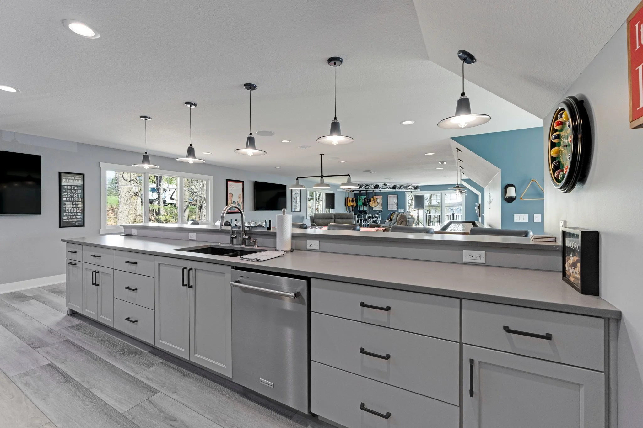 Modern open-concept kitchen with light gray cabinets, white countertop, stainless steel sink, and multiple hanging pendant lights. Large window overlooking trees, with living area in the background featuring guitars on the wall and cozy seating.