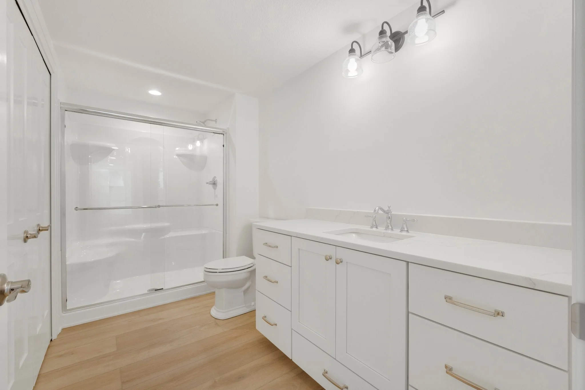 A modern bathroom with a white vanity, marble countertop, and a mirror with four lights. There is a toilet next to the vanity and a glass-enclosed shower with a sliding door. The flooring is light wood.