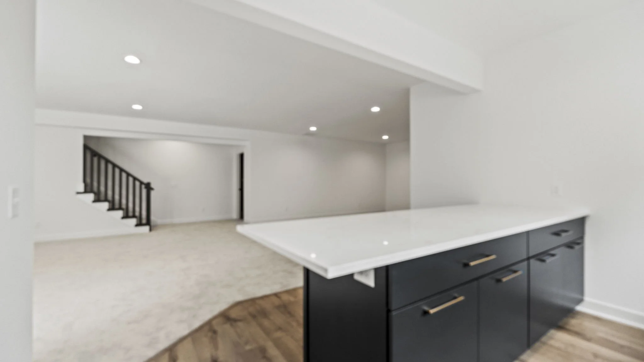 Modern open-concept living space with a kitchen featuring a white countertop and dark cabinets, a stairway with black railing, and white walls and ceiling with recessed lighting.