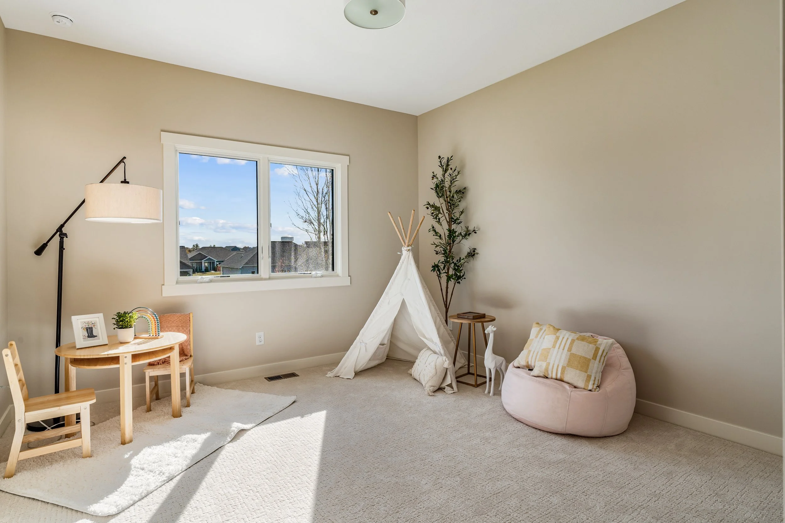 A neutral-toned children's playroom with a window, teepee, plush floor cushion, small wooden furniture, a floor lamp, stuffed giraffe, and decorative plants.