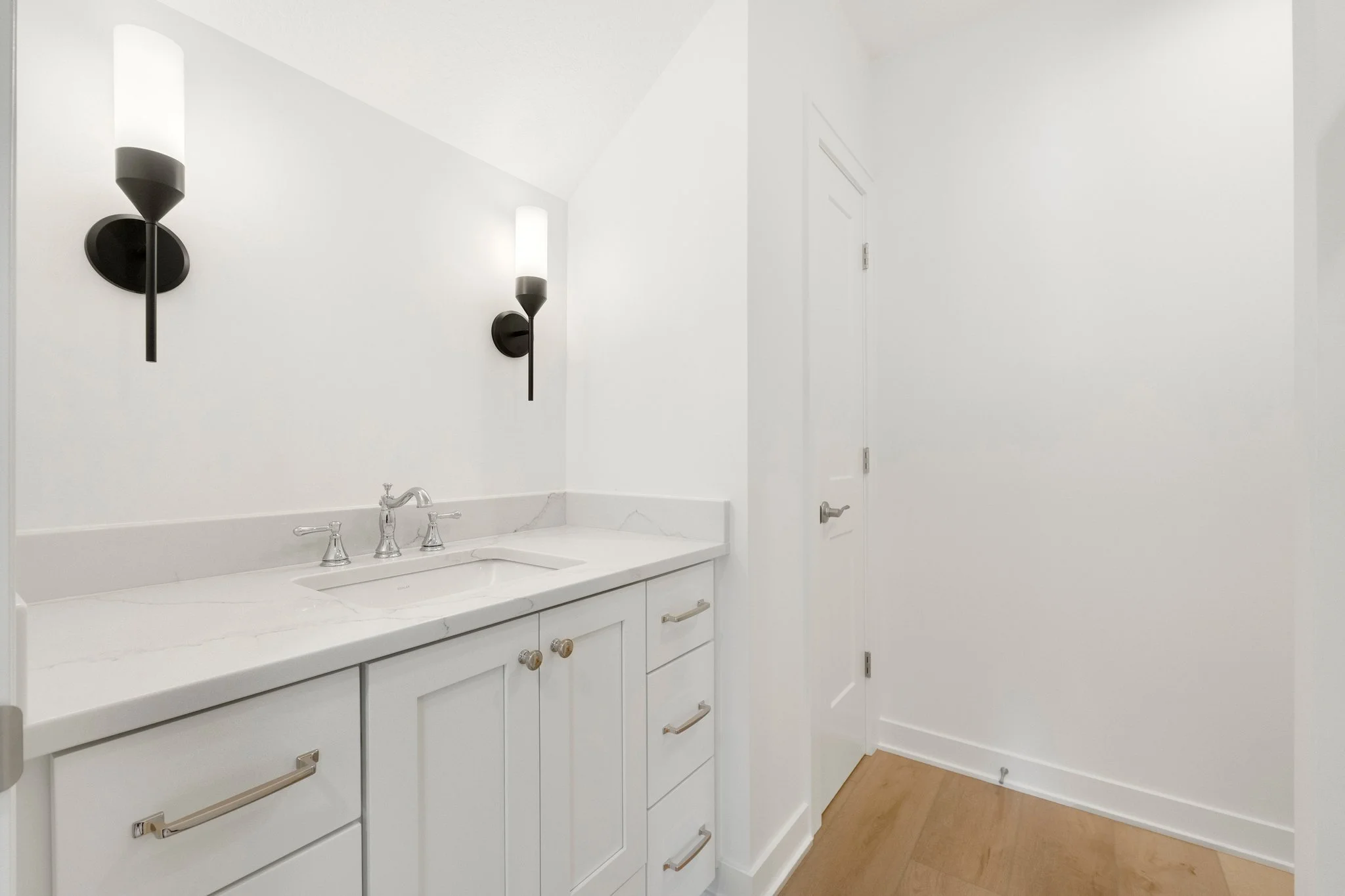 Interior of a modern white bathroom vanity with a marble countertop, two wall-mounted light fixtures, and a white door on the right, with light wood flooring.