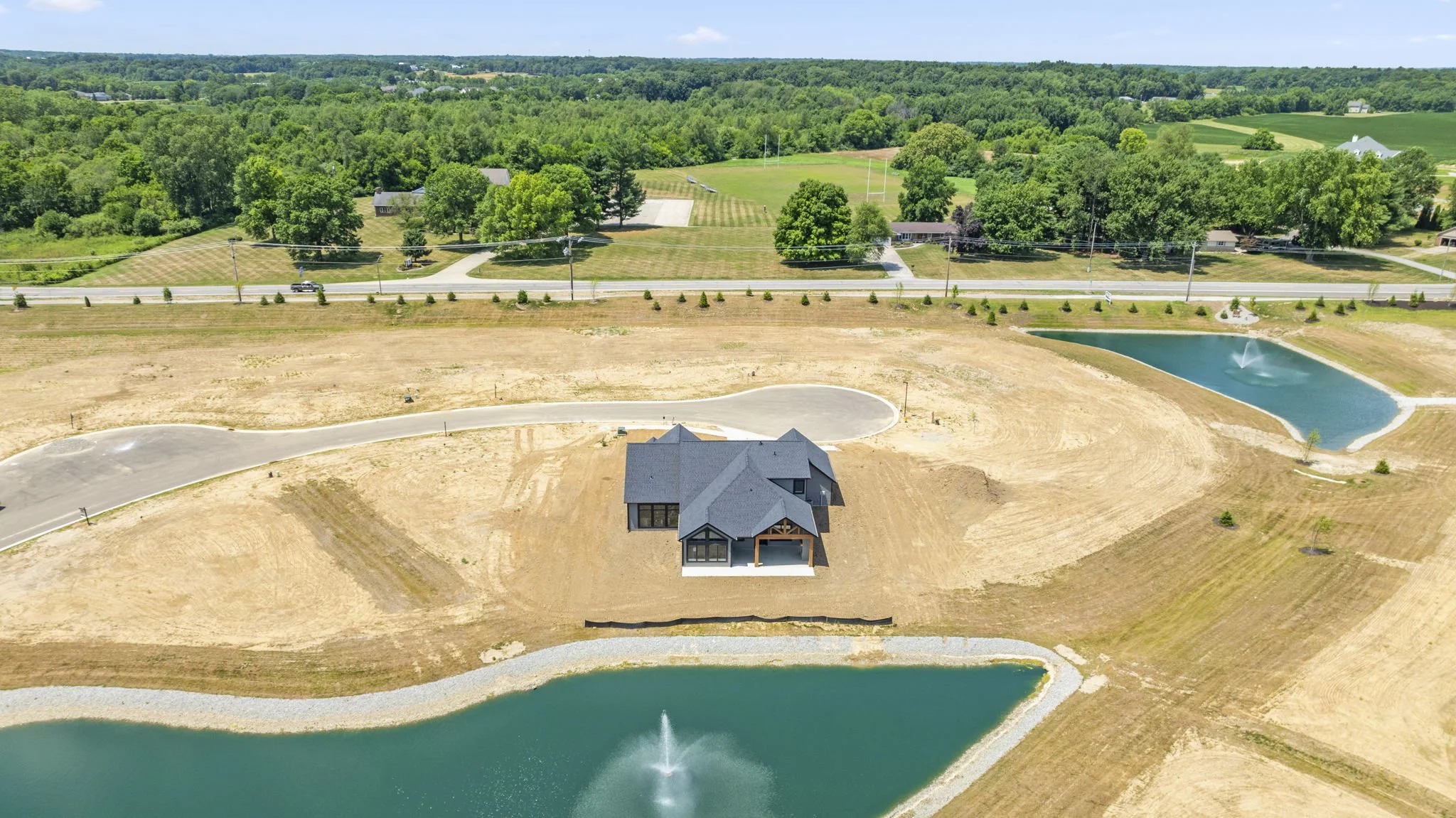 A house under construction in a rural area with two ponds and a fountain, surrounded by dirt and some trees, with a green forest in the background.
