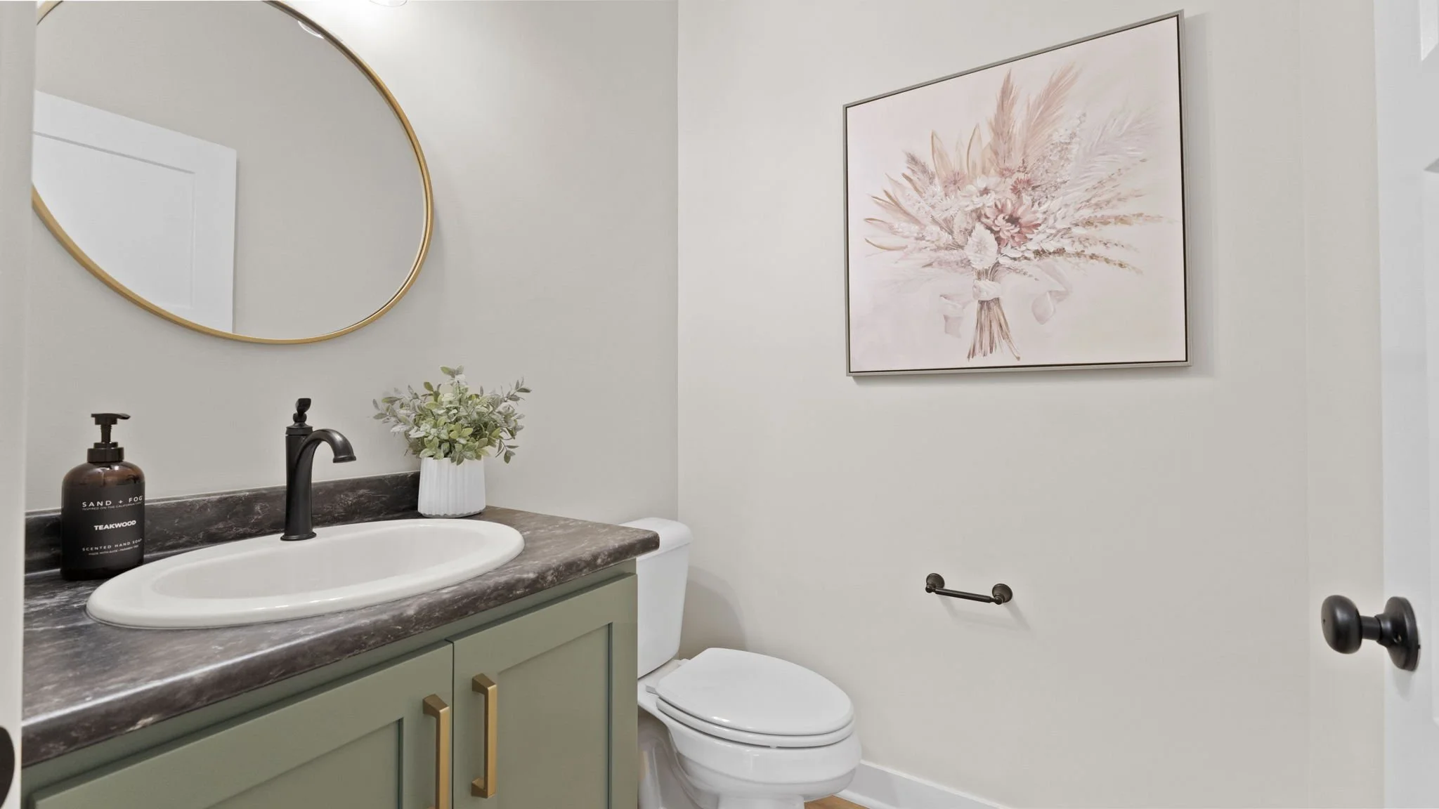 A bathroom with a countertop sink, a black faucet, a dark soap dispenser, a white vase with greenery, a framed pink floral painting on the wall, a mirror with a gold frame, a toilet, and a towel bar.