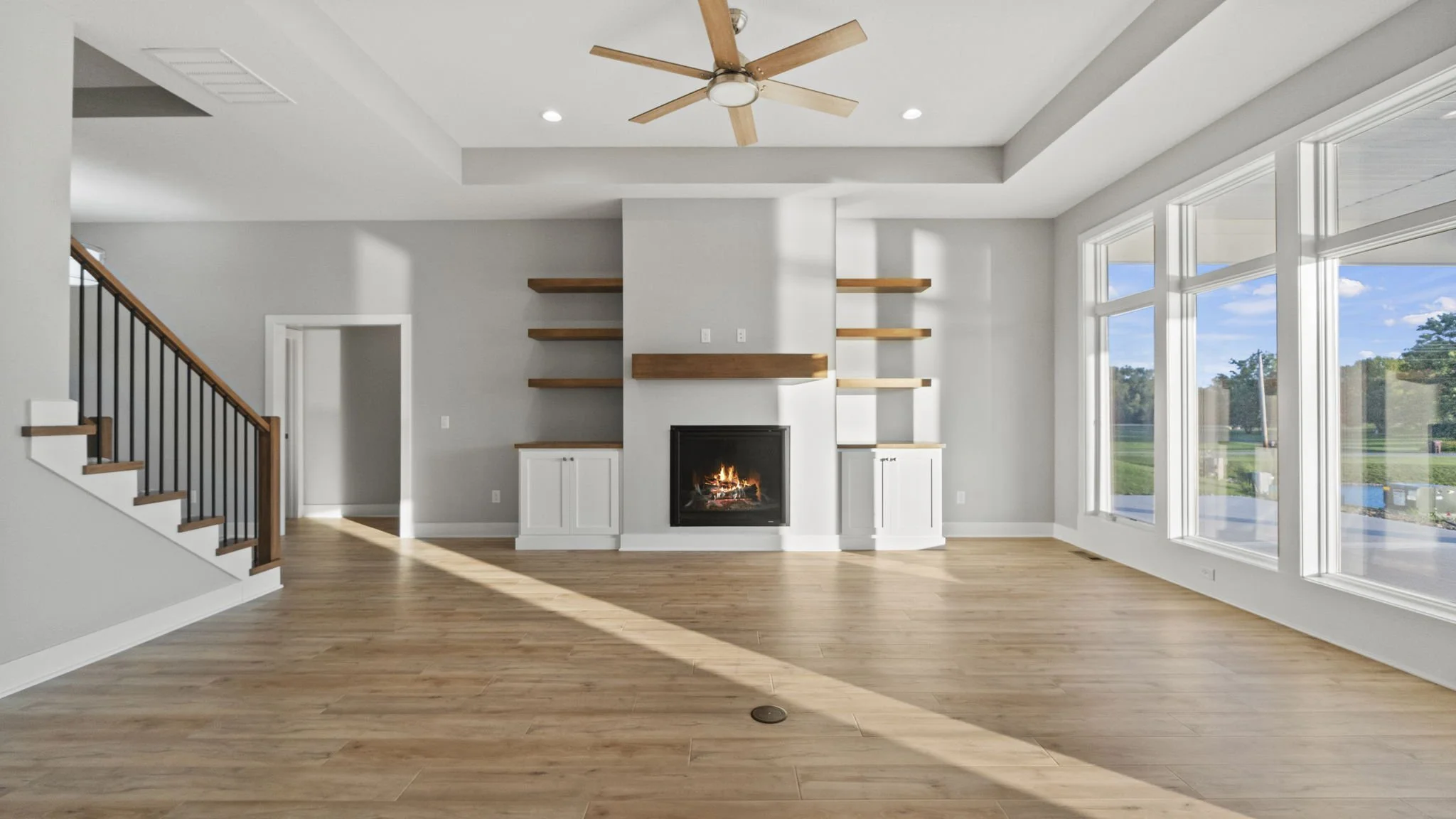 Empty modern living room with large windows, hardwood floors, white cabinetry around a fireplace, built-in wooden shelves, a ceiling fan, and a view of the outdoors.