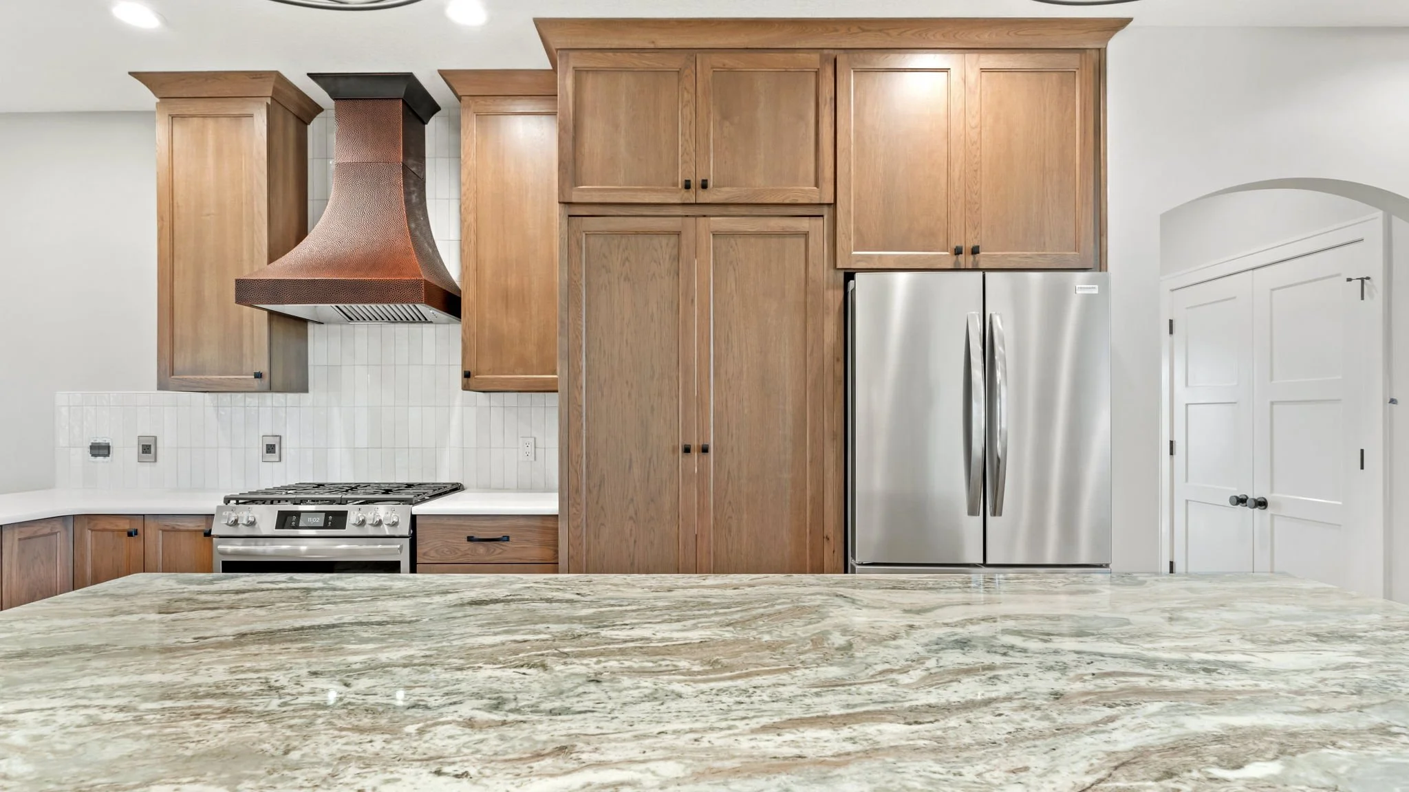 Kitchen with wooden cabinets, stainless steel refrigerator, stove, copper range hood, white tile backsplash, and granite countertop.