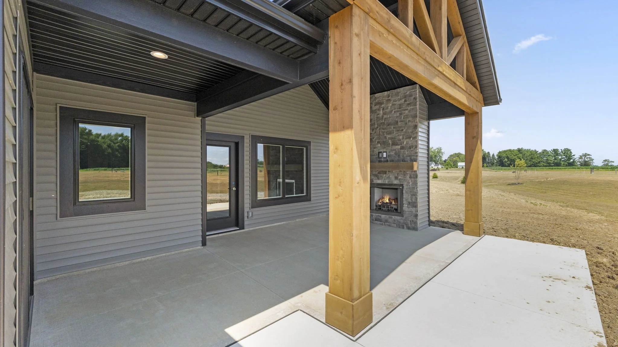 Covered patio area of a house with a fireplace, gray siding, central door, and windows, with a view of an open field in the background.