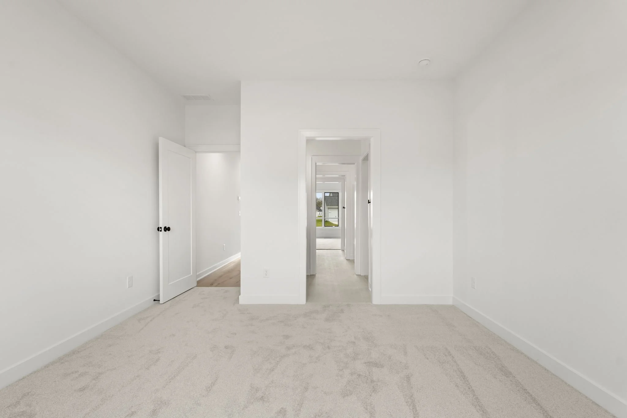 Empty white bedroom with beige carpet, open door to hallway, and window at the end of the hallway showing a house outside.