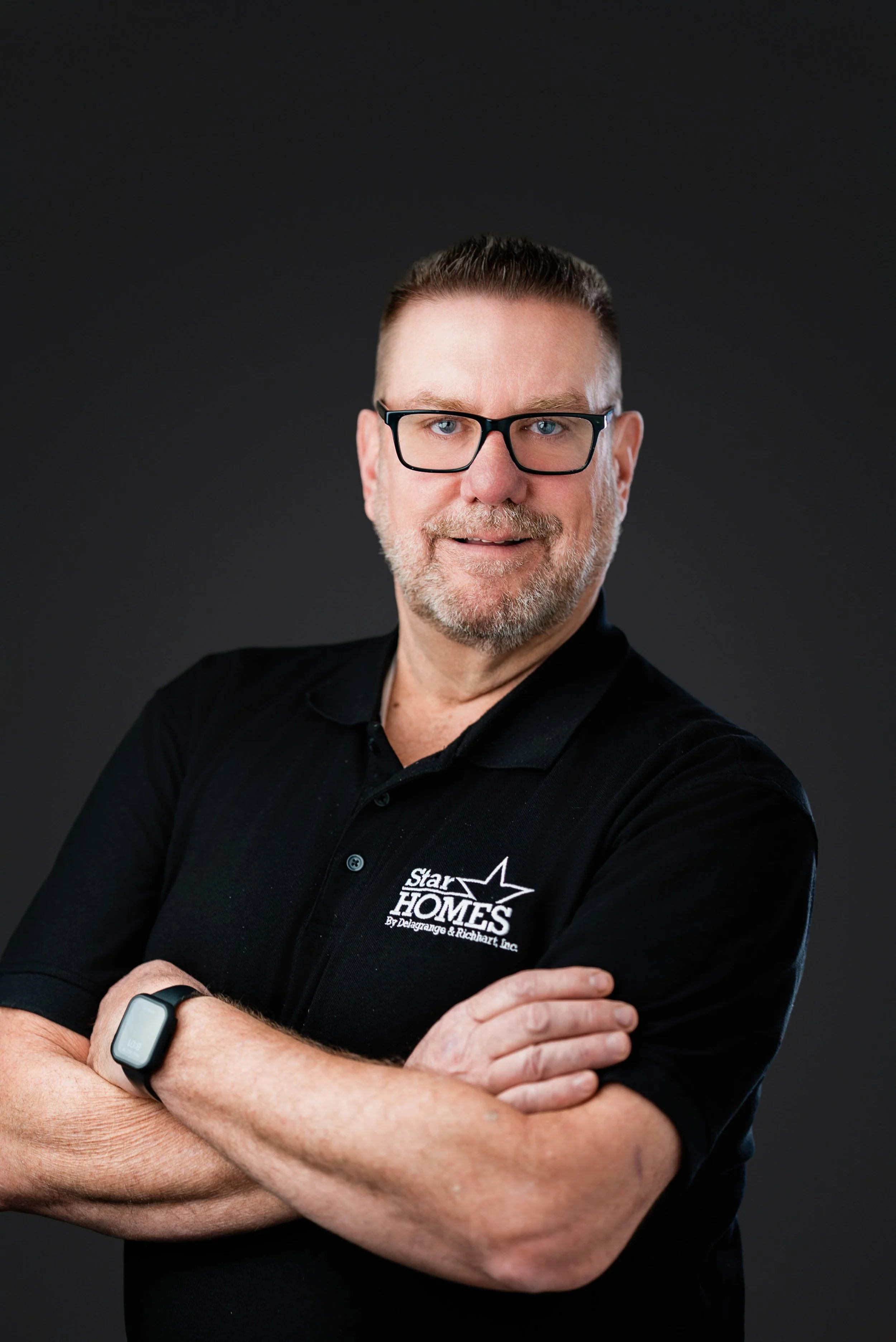 Portrait of a man wearing black glasses and a black polo shirt with a logo, crossing his arms and smiling, against a dark gray background.