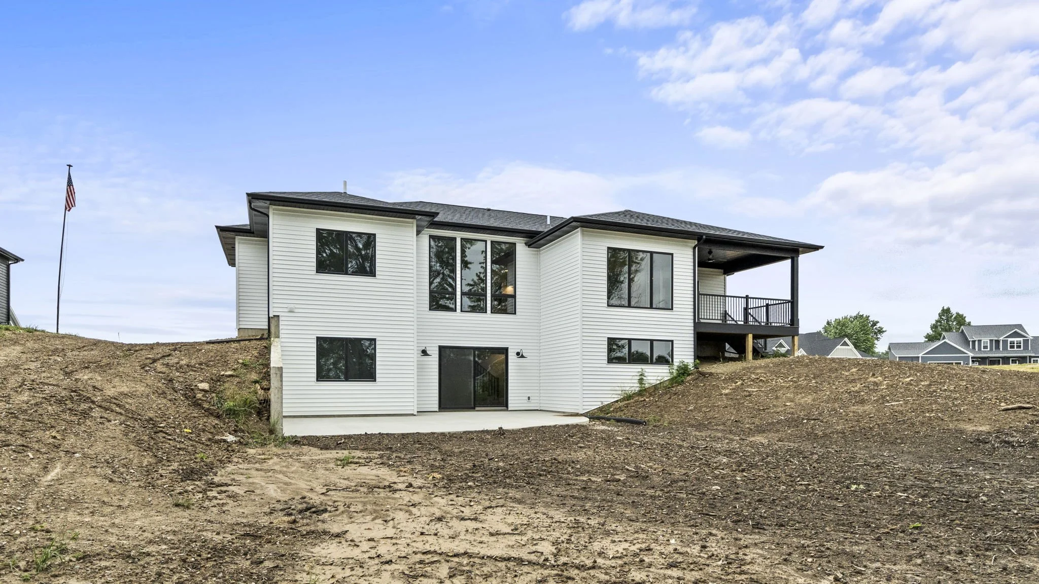 New white two-story house with black roof and black window frames on a lot with dirt and some grass, under a partly cloudy sky.